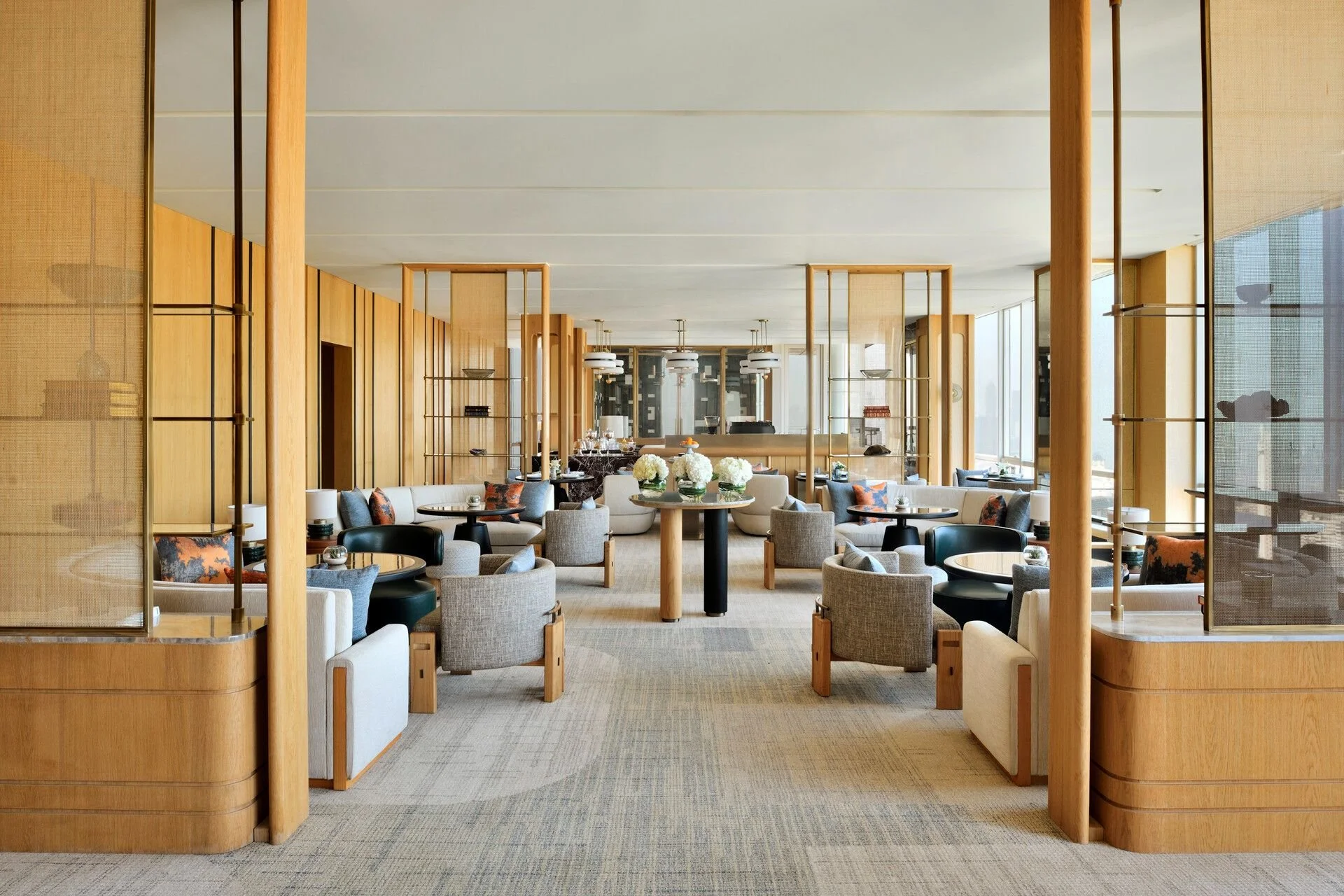 Elegant hotel lounge with neutral-toned furniture, glass flowers, round tables, and decorative wood dividers, with large windows allowing natural light.