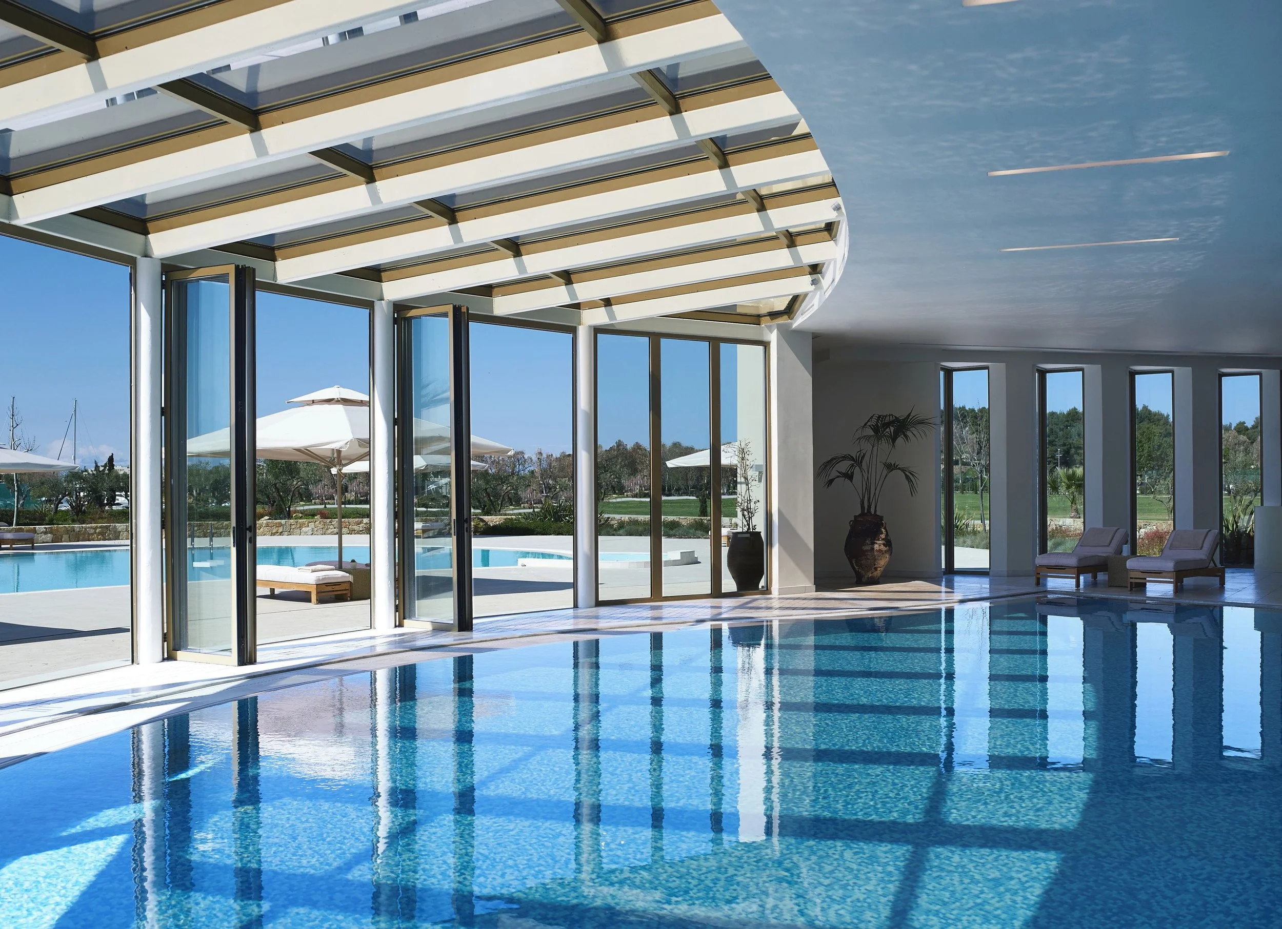 Indoor swimming pool area with large glass walls and ceiling, lounge chairs, potted plants, and a view of outdoor umbrellas and trees.