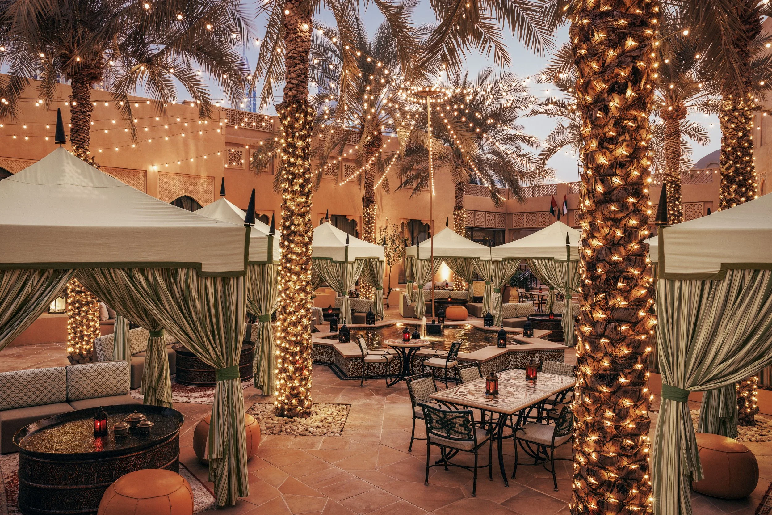 Outdoor patio area decorated with string lights and palm trees wrapped in lights, featuring tents, seating, lanterns, and a fountain, in a warm, inviting evening setting.