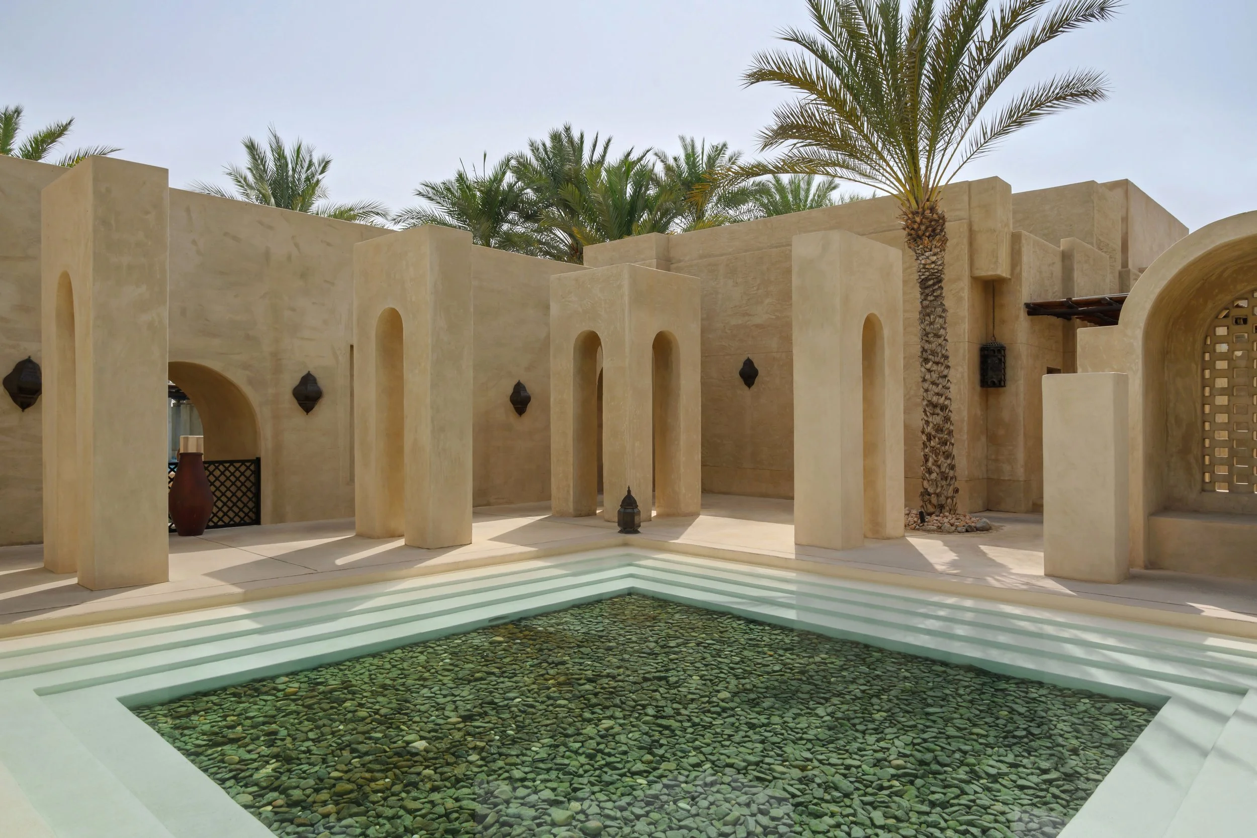 A modern courtyard with beige stucco walls, tall palm trees, and a shallow water feature with green pebbles, set against a cloudy sky.