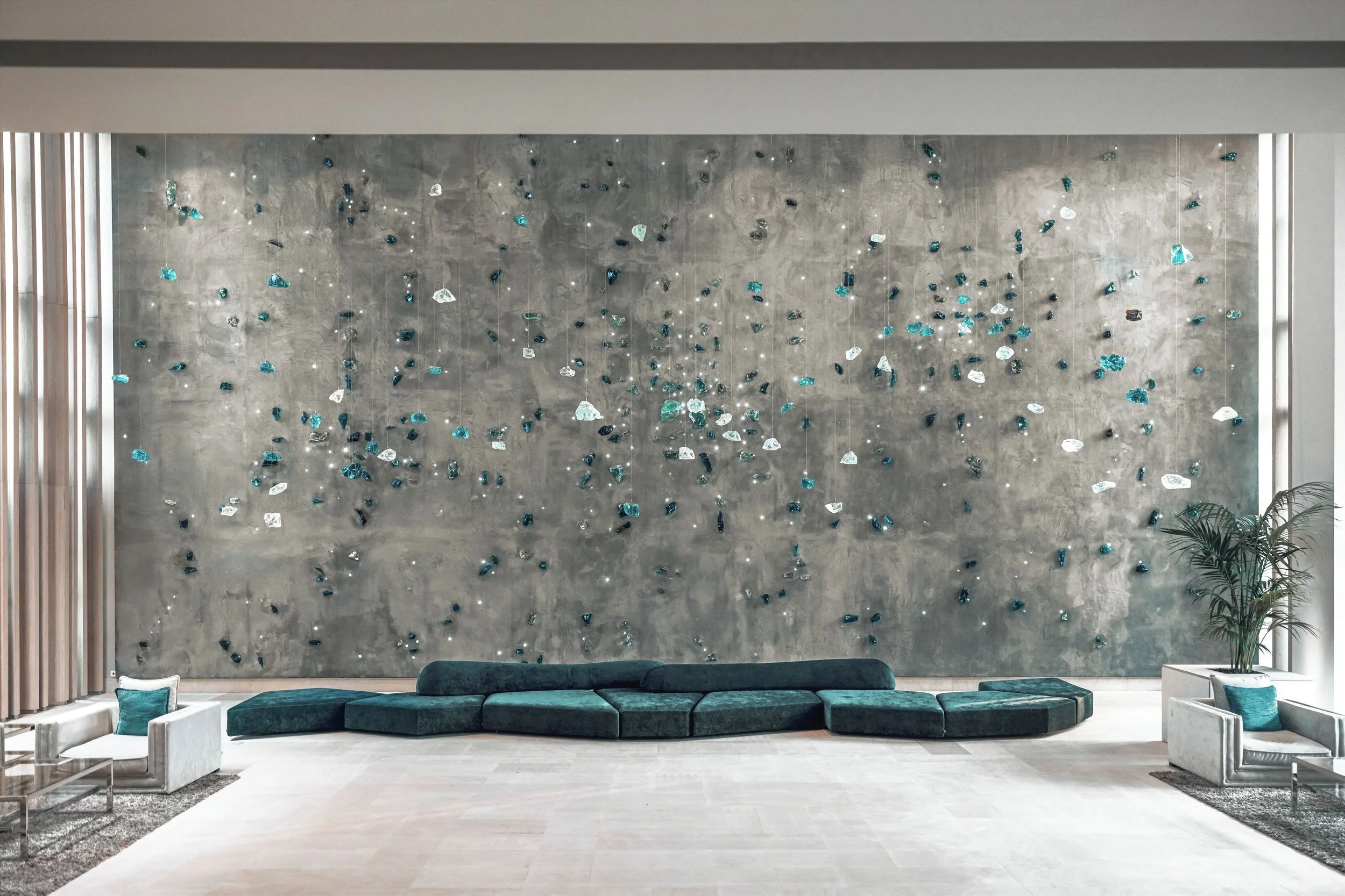 Modern interior with a large textured gray wall decorated with hanging glass art pieces in blue, white, and transparent colors. There is a dark green sofa with a matching ottoman beside it, white chairs with teal cushions, and a tall indoor plant on the right.