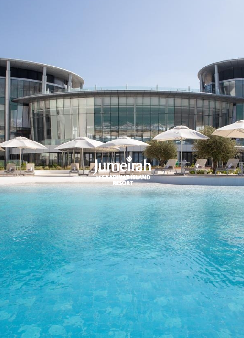 Luxury resort with a swimming pool in the foreground, poolside lounge chairs and umbrellas, and a modern glass building behind, featuring a clear sky.
