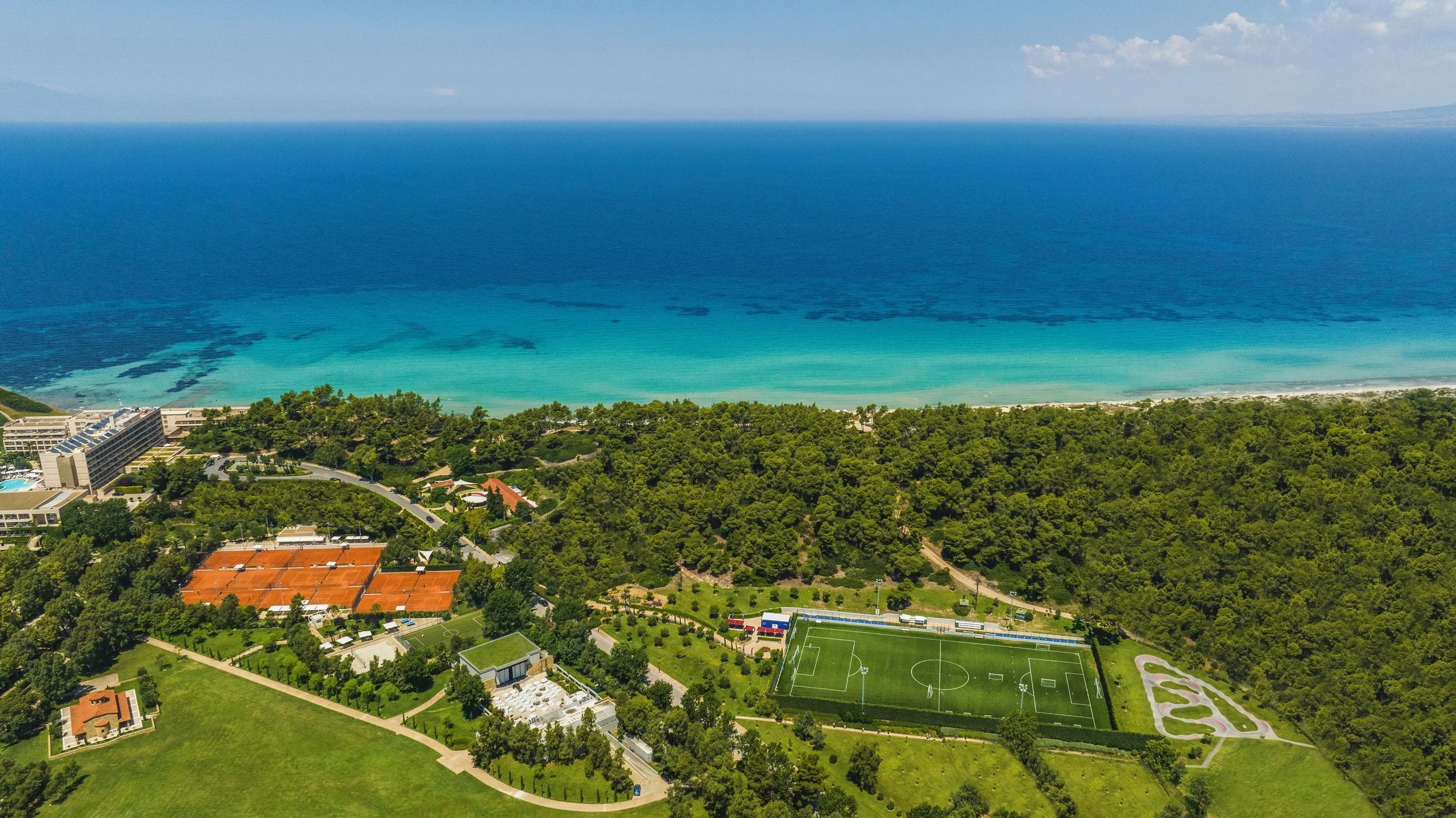 Aerial view of a coastal area showing a large body of blue ocean waters meeting a beach with some green vegetation. There are buildings, sports fields, and roads along the shoreline.