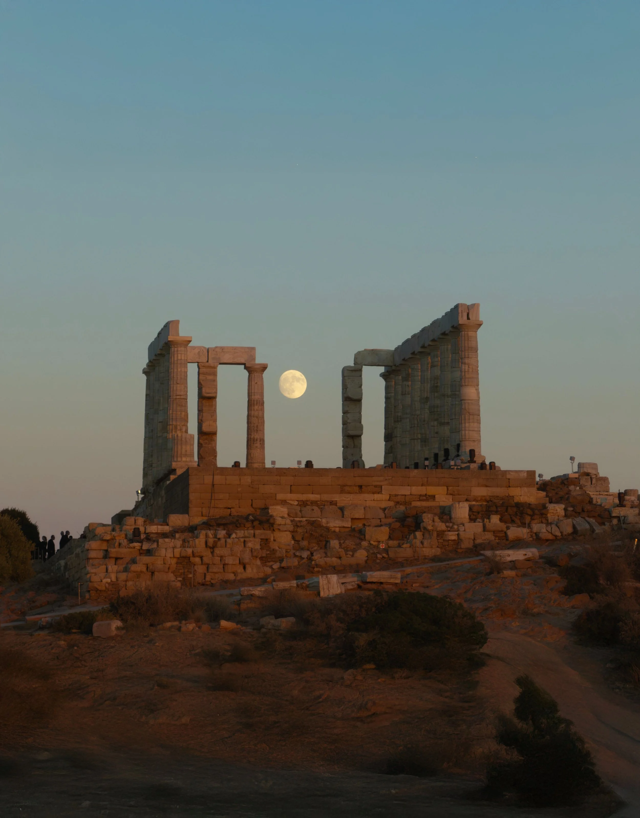 The moon is rising behind the ancient ruins of an ancient Greek or Roman temple, with large stone columns and a stone platform in the foreground. The sky is clear, showing the early evening or twilight setting.