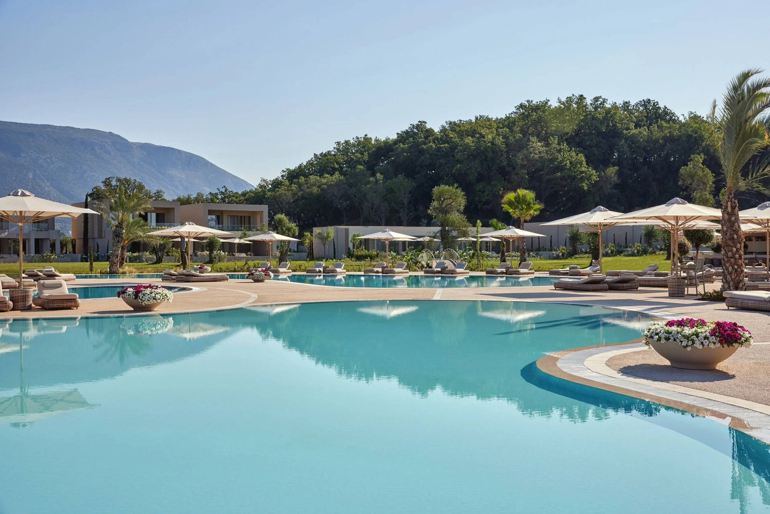 Luxury outdoor pool area with lounge chairs, umbrellas, potted plants, green trees, apartment buildings in the background, mountains, and a clear, blue sky.