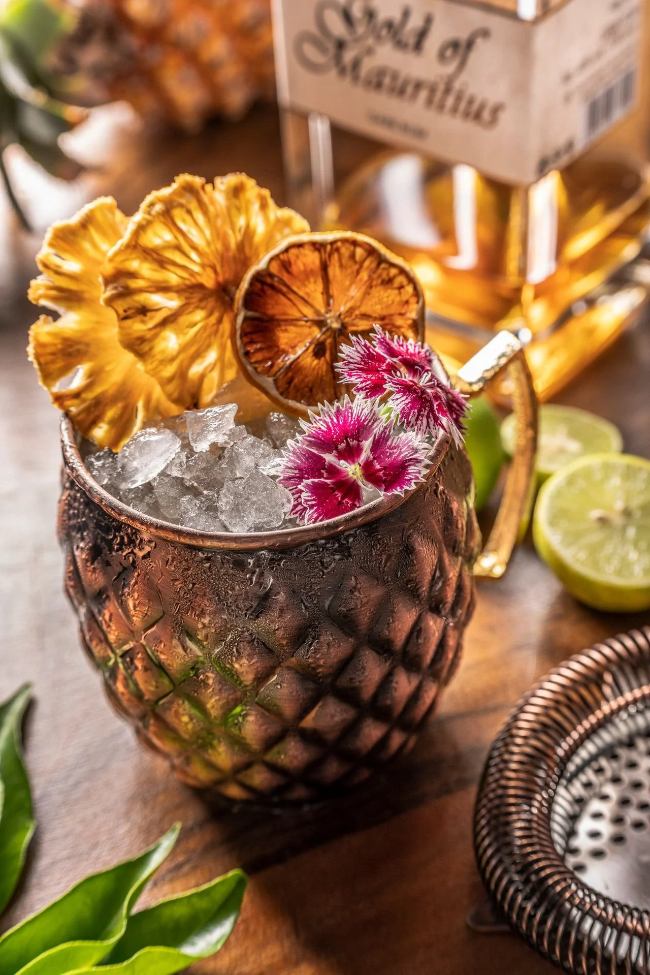 A decorative pineapple-shaped container filled with ice, garnished with dried pineapple and citrus slices, and pink carnations, on a wooden surface with a blurred background.