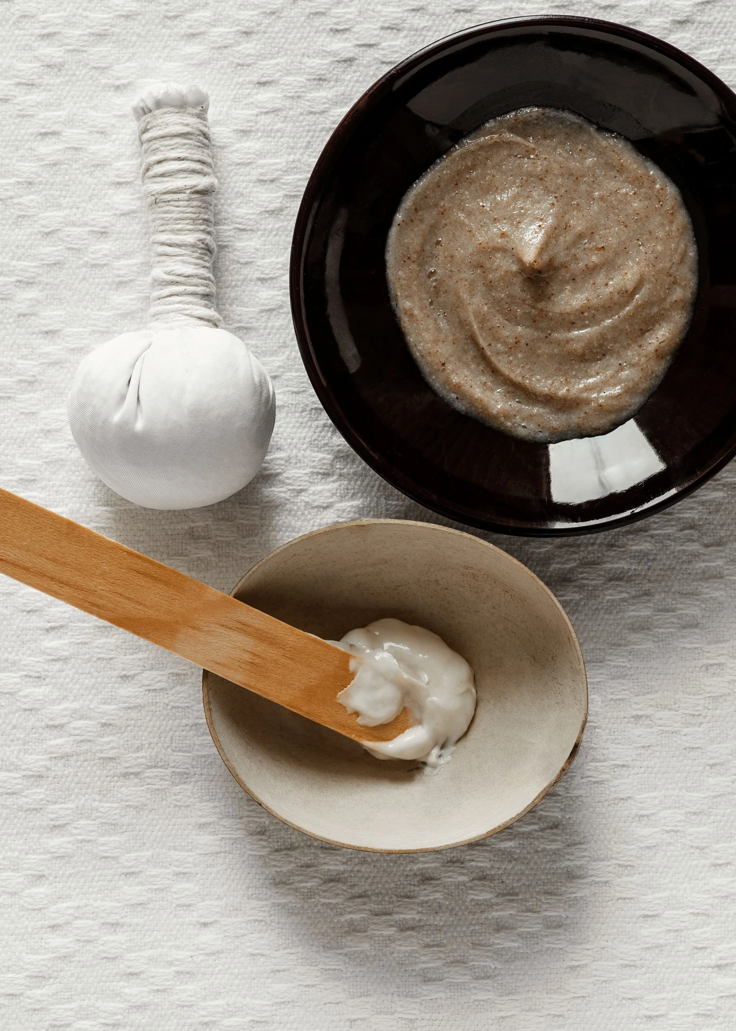 A black bowl containing a creamy, textured substance, next to a white fabric pouch with a wrapping tied at the top, and a wooden utensil with a dollop of white creamy material inside a small beige bowl on a white textured surface.