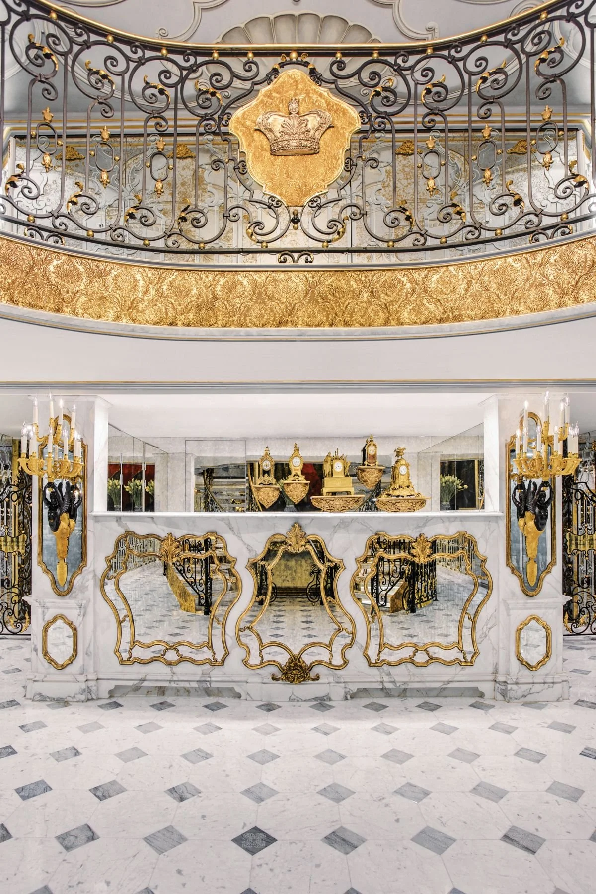 Luxurious hotel lobby with ornate gold and white decor, featuring a marble floor, elaborate wrought iron balcony with gold accents, and a marble reception desk decorated with gold details. The background shows a collection of antique clocks and a mirror.
