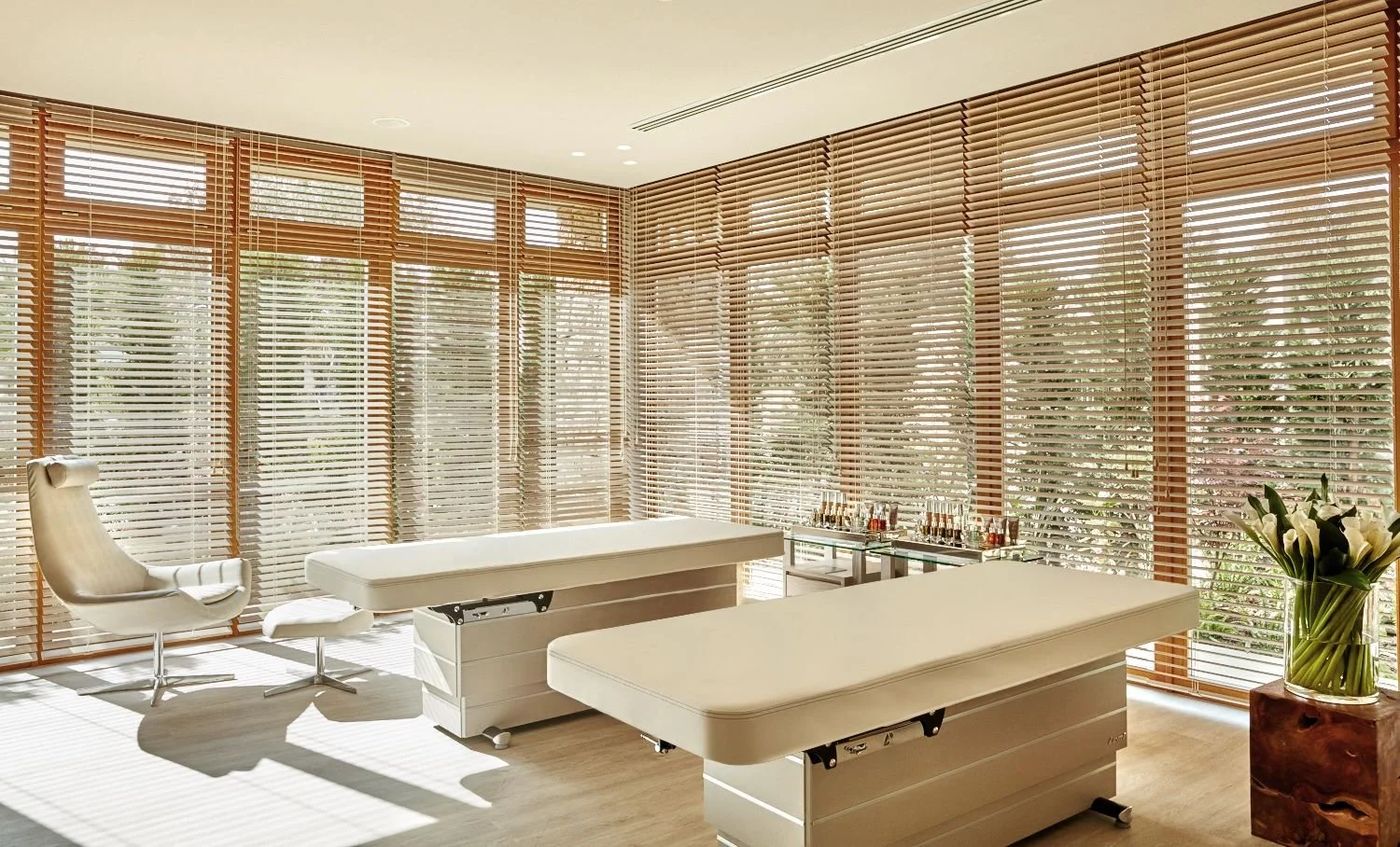 A bright, modern medical or spa treatment room with large windows covered by wooden blinds. There are two treatment tables, a leather chair with an ottoman, and a glass table with bottles. A vase of white lilies is on a wooden stand.