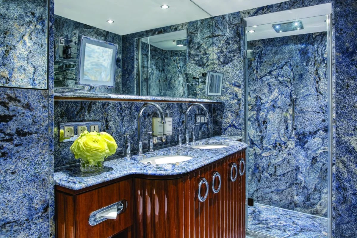 A luxurious bathroom with blue and gray marble walls and countertops, a wooden vanity with three sinks, a large mirror, a vase with yellow flowers, and a shower area with a glass door.