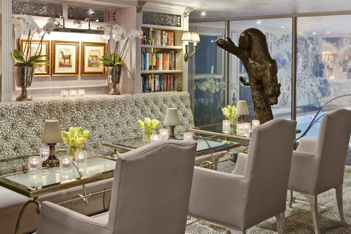 Elegant dining area with glass-topped tables, white upholstered chairs, decorative lamps, floral arrangements, a sculpture of a horse's head, and a bookshelf in the background.