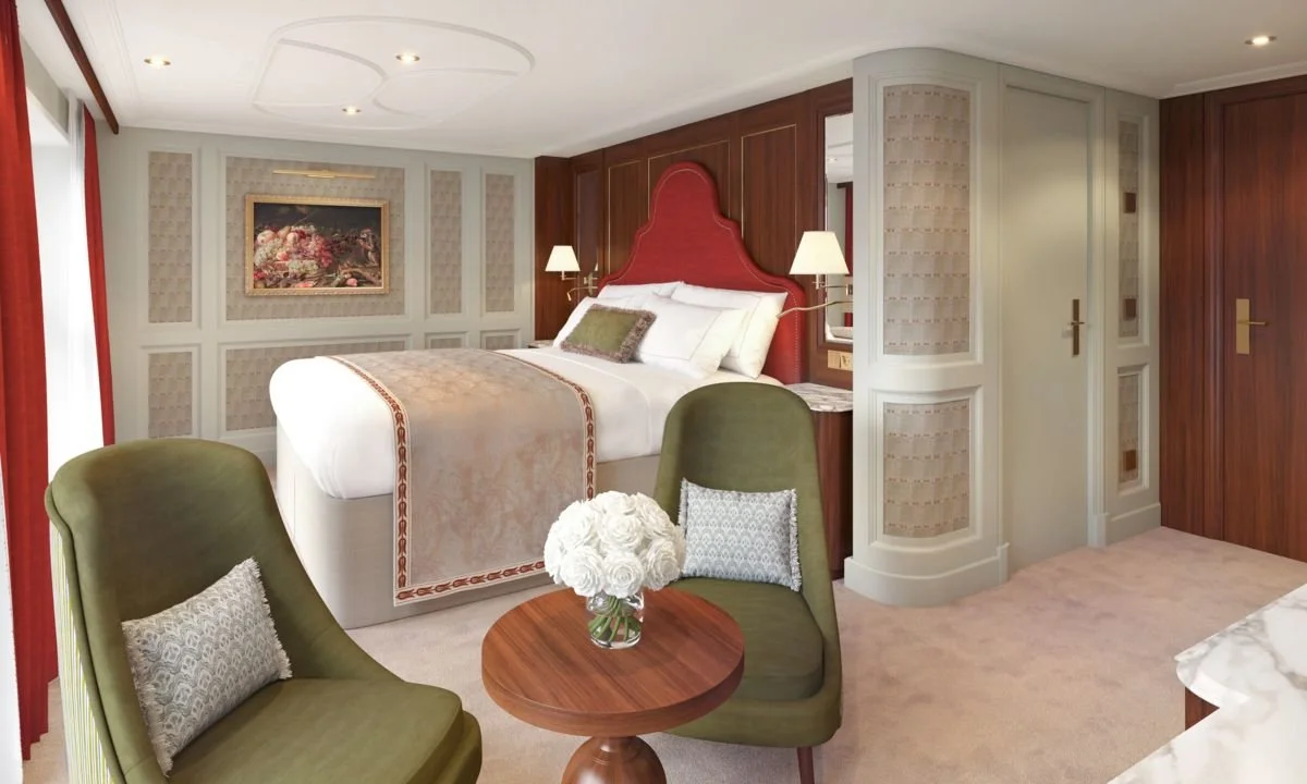 Elegant hotel room featuring a bed with a red headboard, bedside lamps, two green chairs with decorative pillows, a small round wooden table with a vase of white flowers, and a classic art piece on the wall.