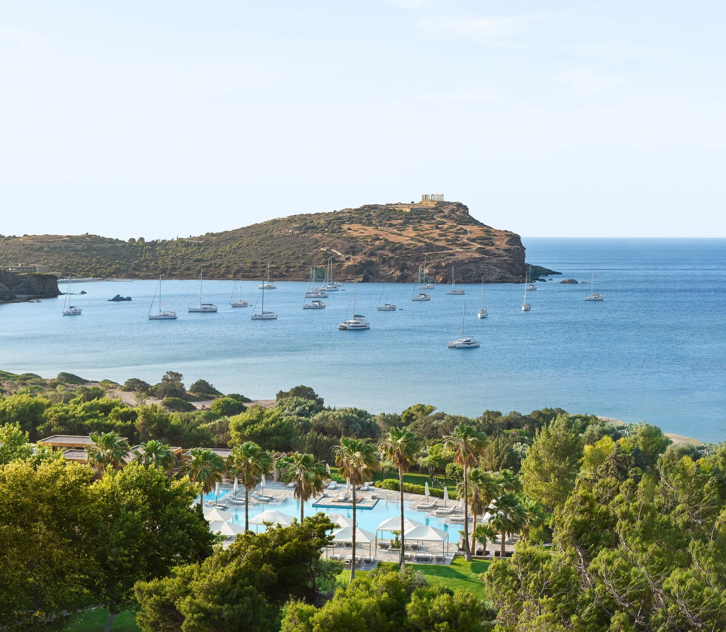 A scenic view of a coastal area with a hill in the background, featuring some ruins or structures on top. There are boats anchored in the water and a lush area with green trees and a swimming pool with sun umbrellas and lounge chairs in the foreground.