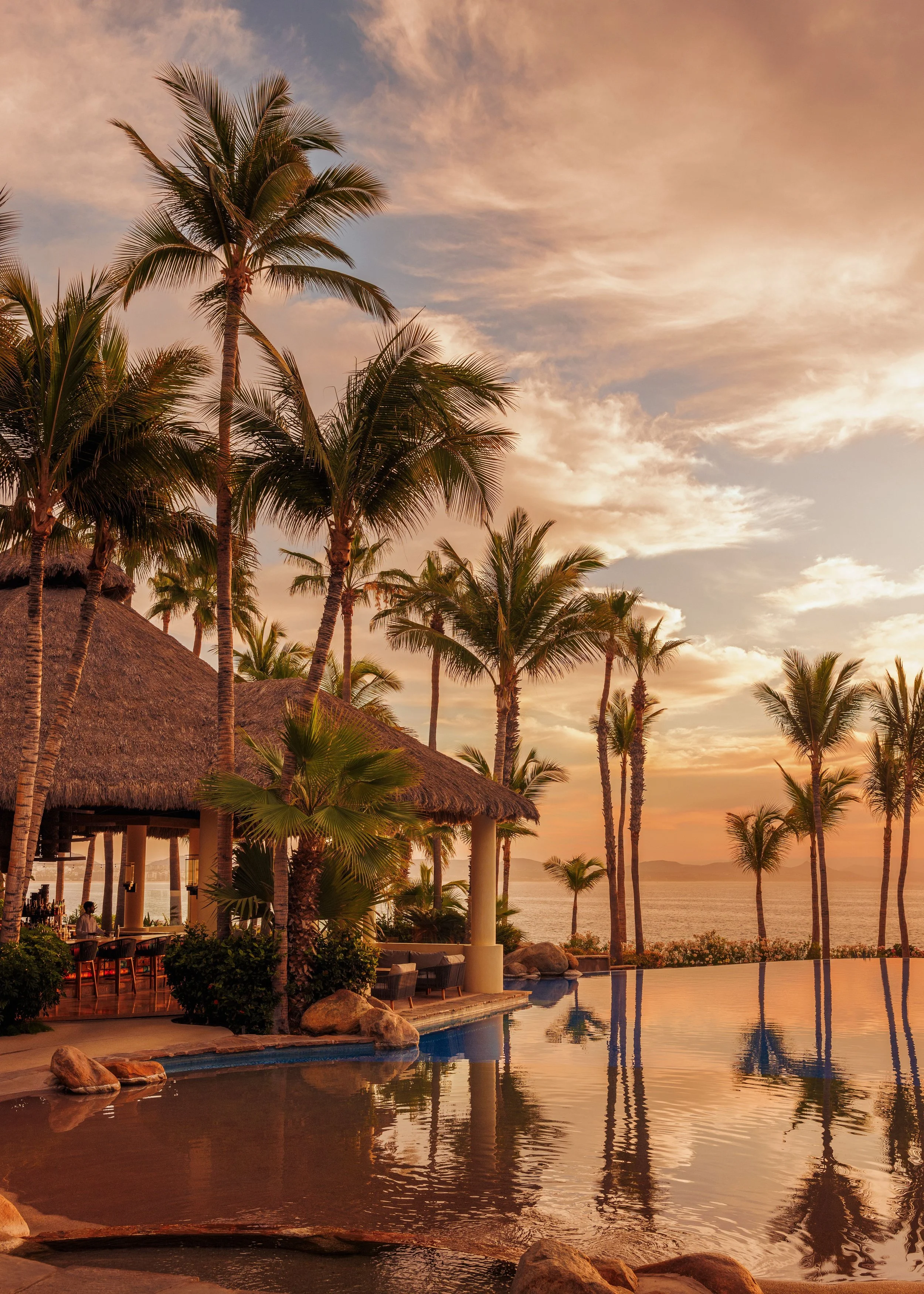 A tropical resort with tall palm trees, a thatched roof hut, a pool, and an ocean view at sunset.