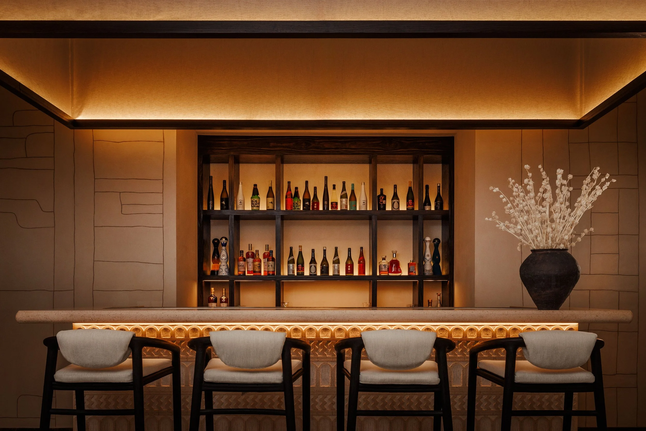 A bar counter with a backlit shelf displaying liquor bottles, a decorative vase with dried flowers, and chairs in front of the counter.