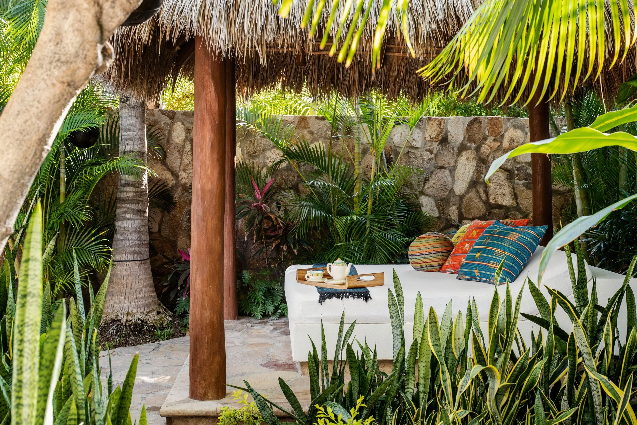 Tropical outdoor seating area with a thatched roof, stone wall, lush green plants, and colorful pillows on a white sofa with a tray of tea cups.