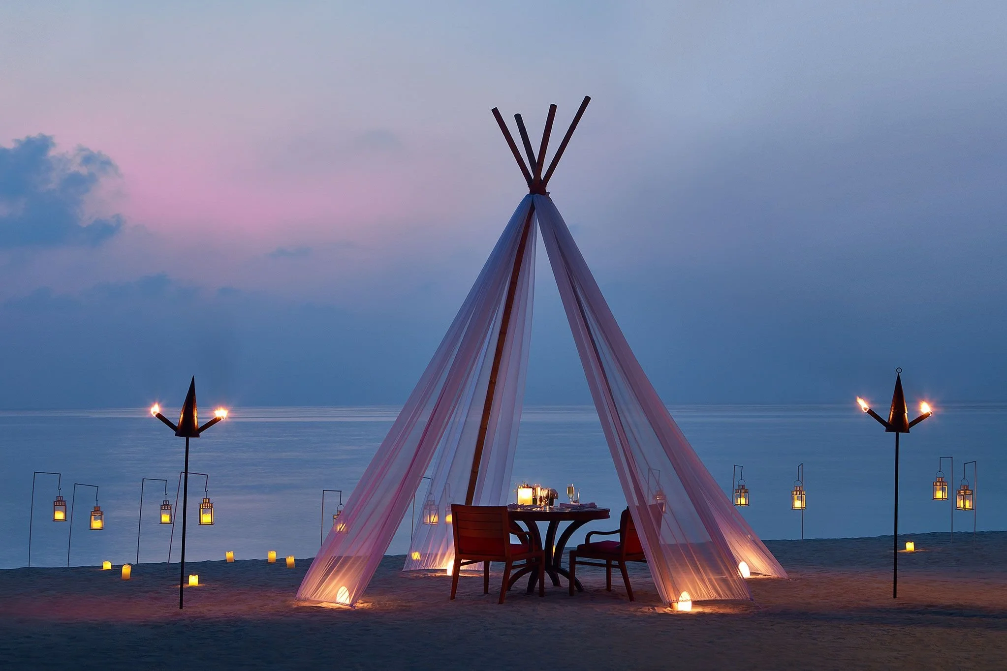 A decorative teepee-like structure outlined with fabric at the beach during sunset, with candles on the ground and lanterns hanging around, against the backdrop of the ocean and a twilight sky.