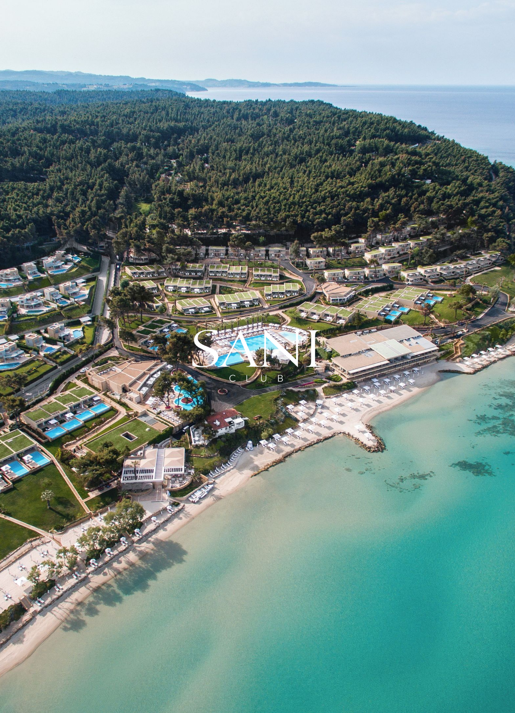 Aerial view of a beachfront resort with pools, white buildings, lush green trees, and a sandy beach along clear turquoise water.