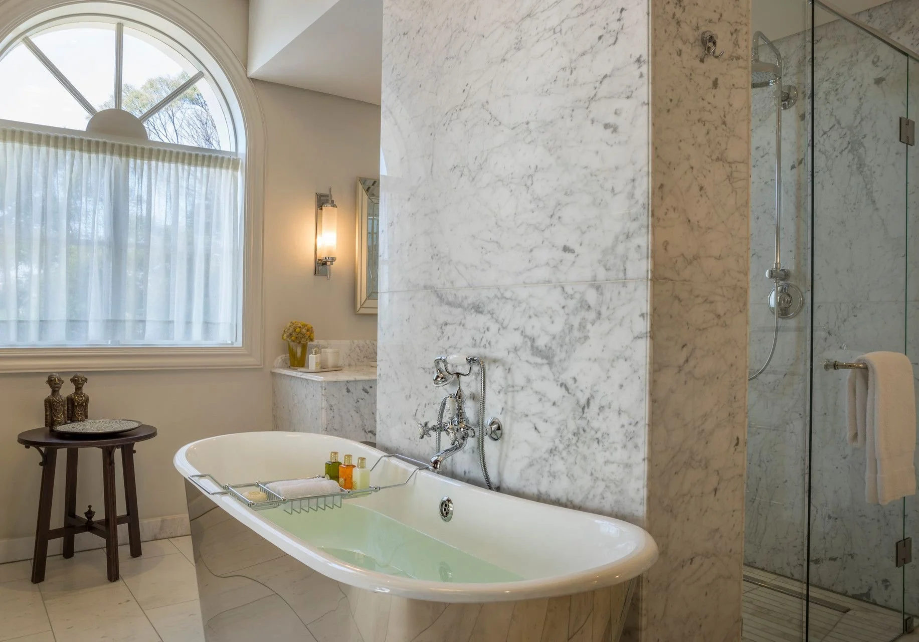 A luxurious bathroom with a freestanding bathtub filled with water, situated in front of a large arched window with sheer curtains. To the right, there is a marble wall with a shower area enclosed by glass. Decor items include a small side table with sculptures, a flower vase, and bath amenities on the bathtub's edge. The space features elegant decor and natural light.
