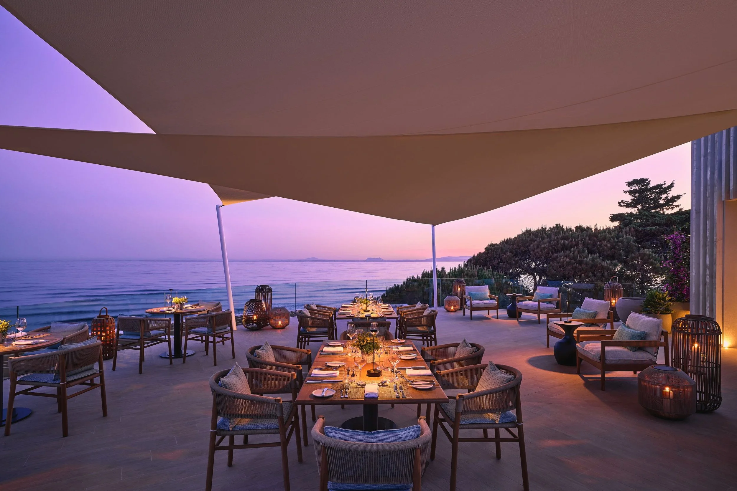 An outdoor restaurant with tables set for dinner, overlooking the ocean at sunset, with purple and pink sky hues. There are cushioned chairs, candles, and decorative lanterns under a large fabric canopy, surrounded by greenery and trees.