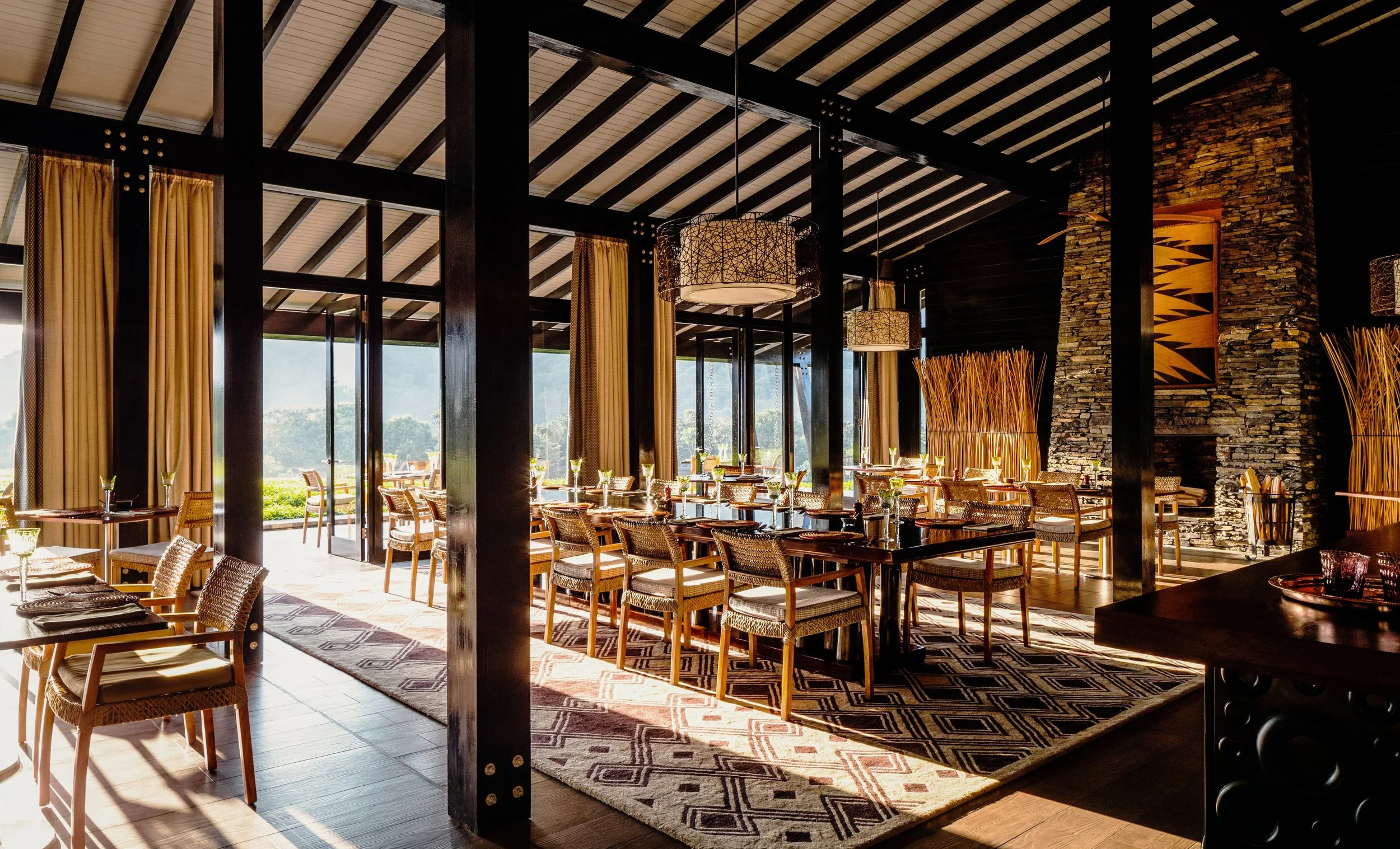 Interior of a stylish restaurant with large windows, wooden furniture, hanging lights, and decorative elements, with outdoor greenery visible outside.
