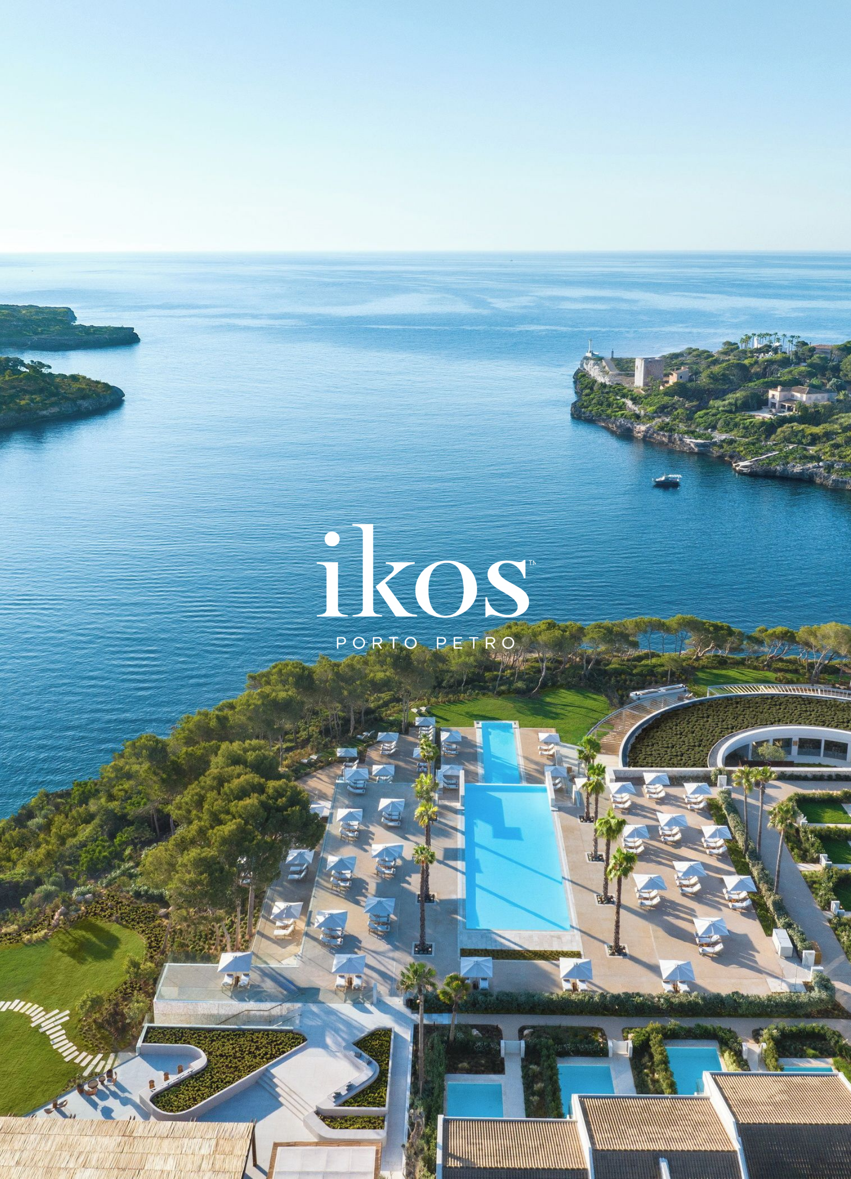 Aerial view of Ikos Porto Petro resort with a large infinity pool, sun loungers, palm trees, and scenic coastal landscape with cliffs and ocean in the background.