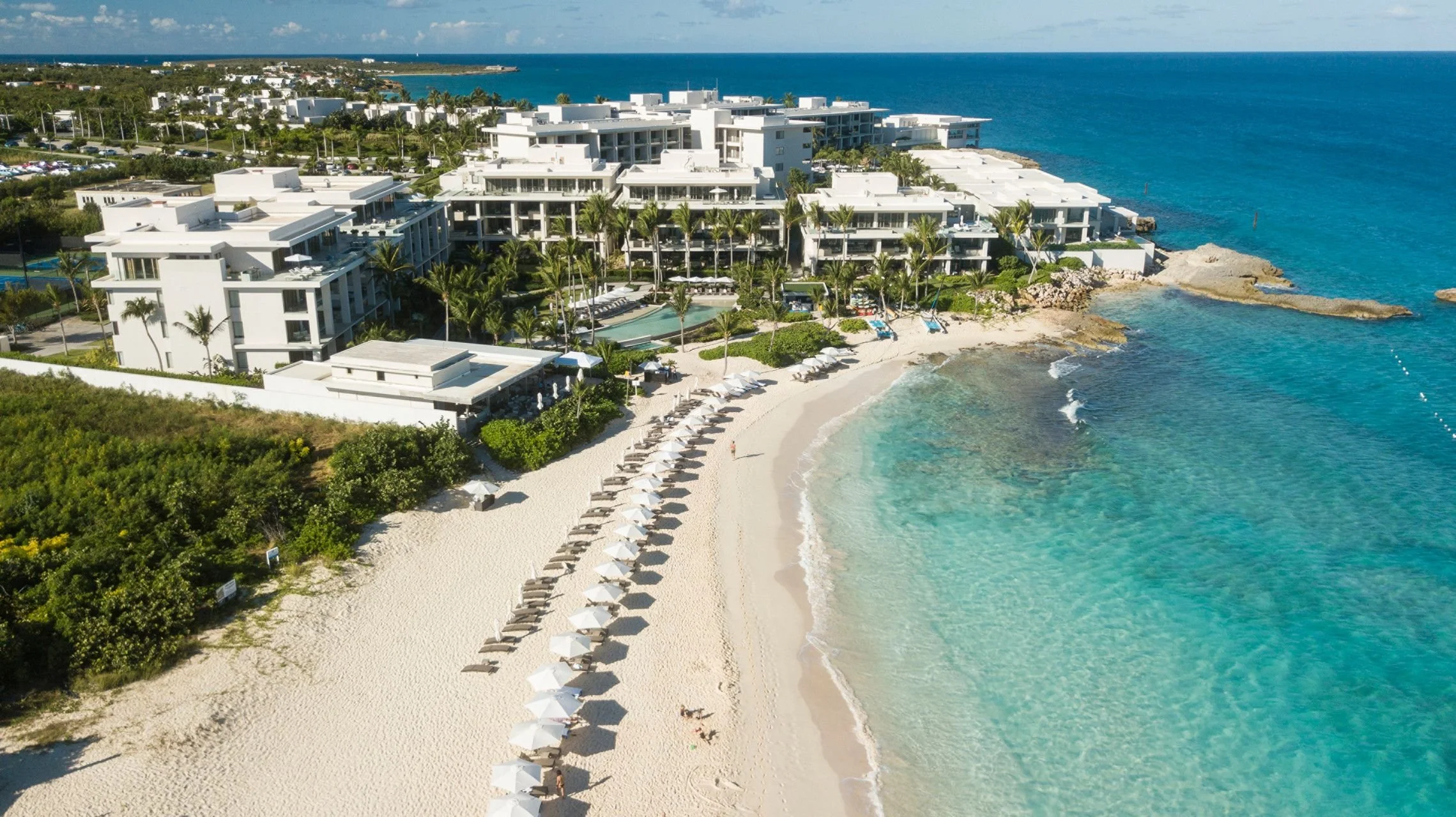 An aerial view of a modern white beachfront resort with multiple buildings, surrounded by palm trees, overlooking clear blue waters with a sandy beach and lounge chairs arranged along the shore.