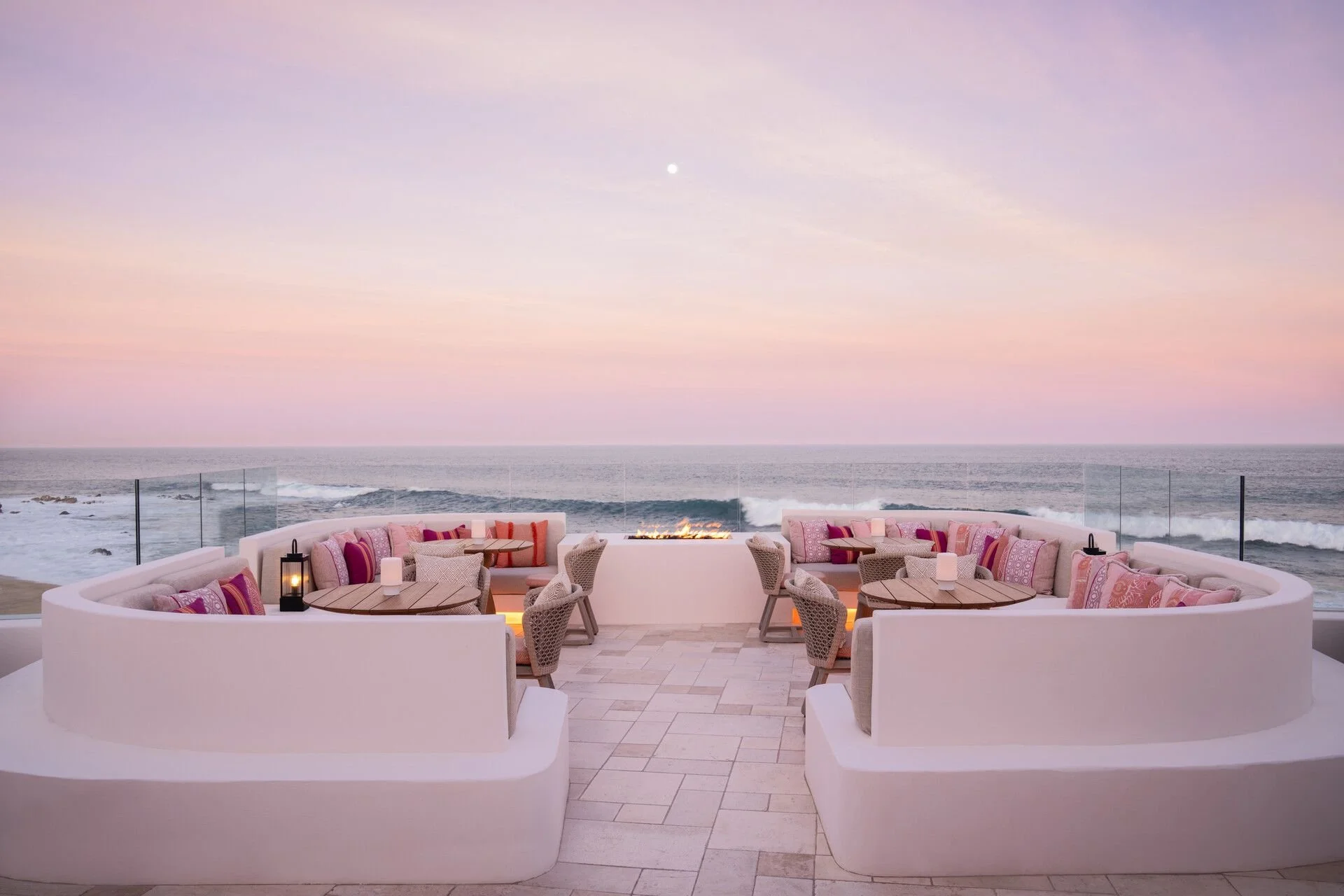 Luxury outdoor seating area with curved white sofas and pink patterned cushions overlooking the ocean during sunset, with a glass barrier, a fire pit, and a pastel sky.