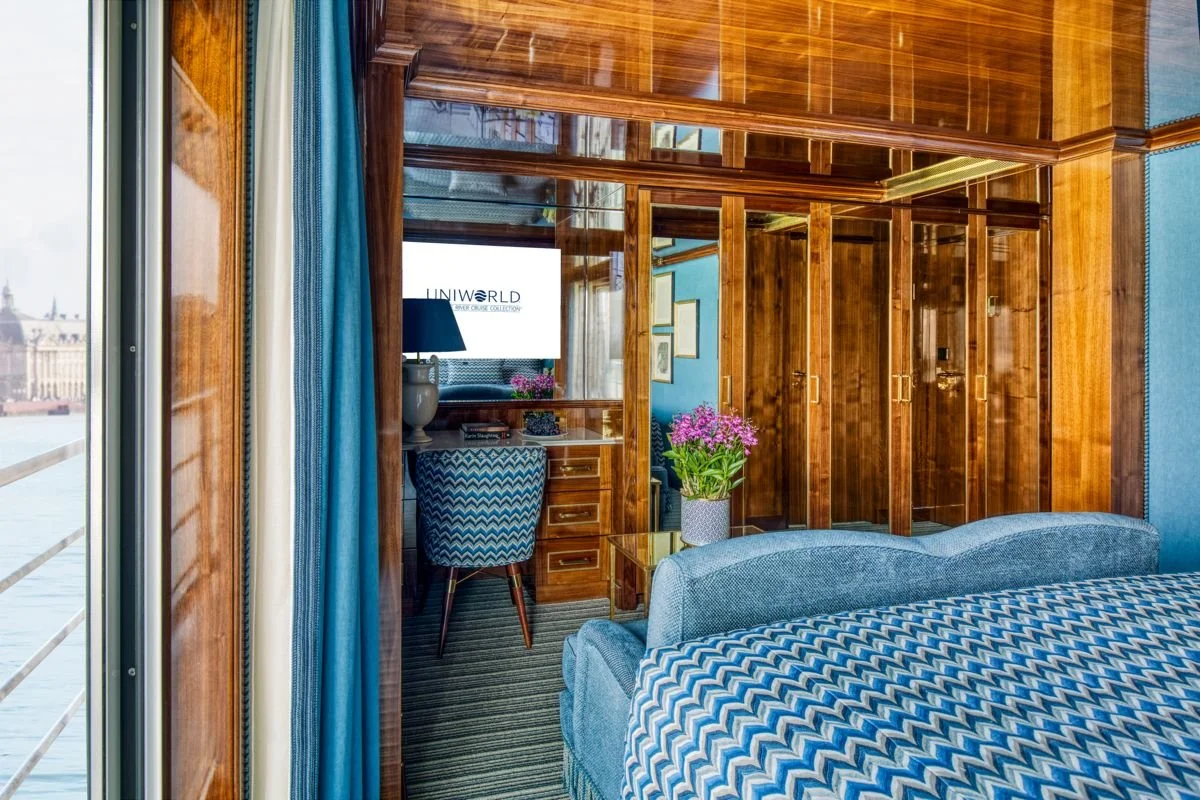 A hotel room with wooden wall partitions, a blue bed with a chevron-patterned bedspread, a window with blue curtains, a vase of pink flowers on a nightstand, a small desk with a blue patterned chair, and a view of a cityscape through the window.