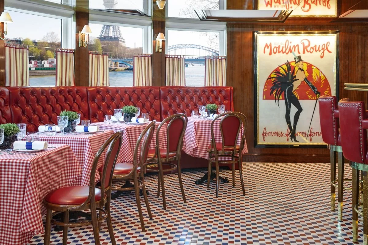 Interior view of a French-themed restaurant with red and white checkered tablecloths, red leather booths, wooden chairs, and Paris-themed decor including a large Moulin Rouge poster. Large windows reveal a river and the Eiffel Tower in the background.