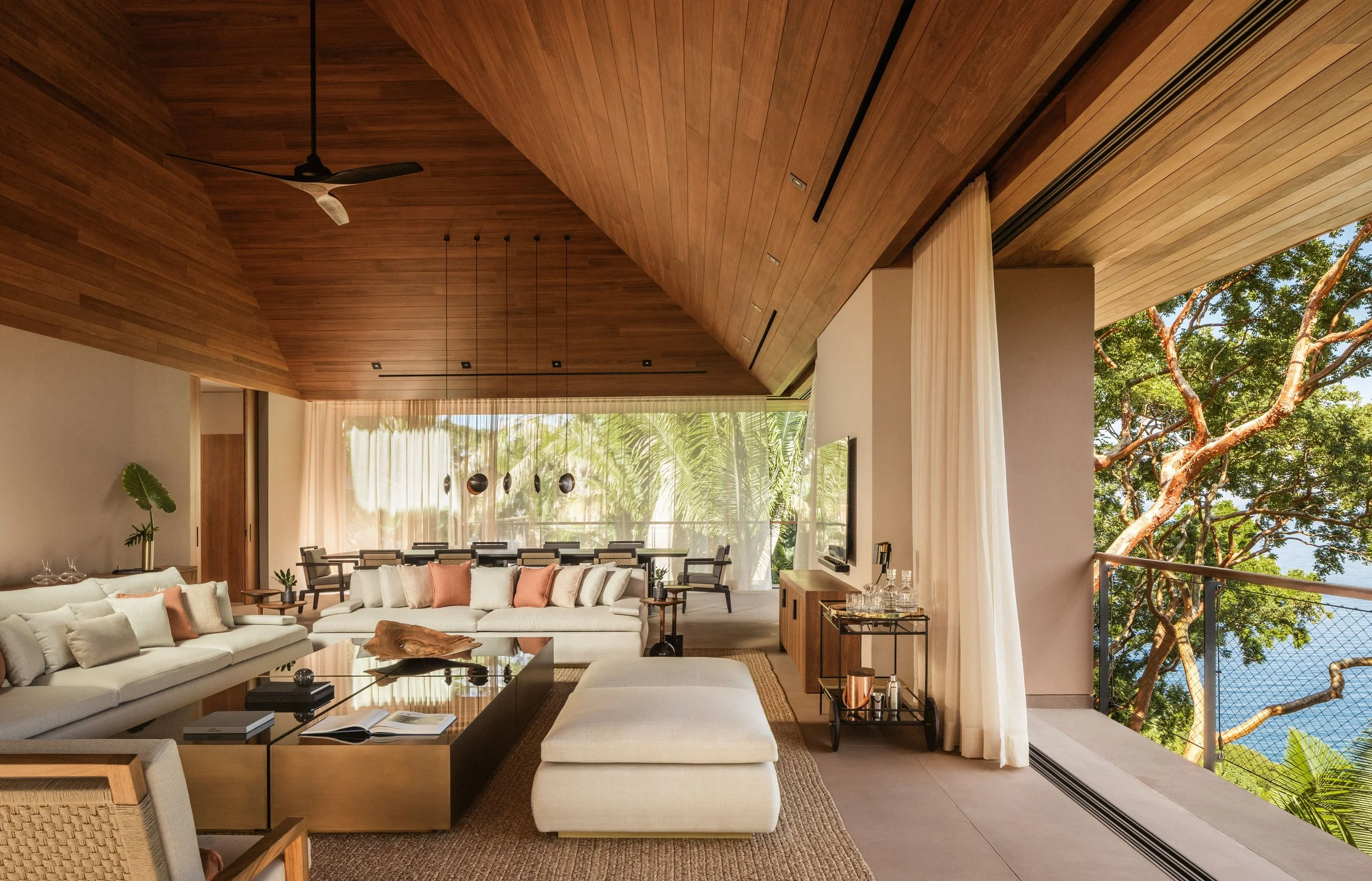 Modern living room with white sofas, a wooden ceiling, large windows, and a balcony with trees visible outside.