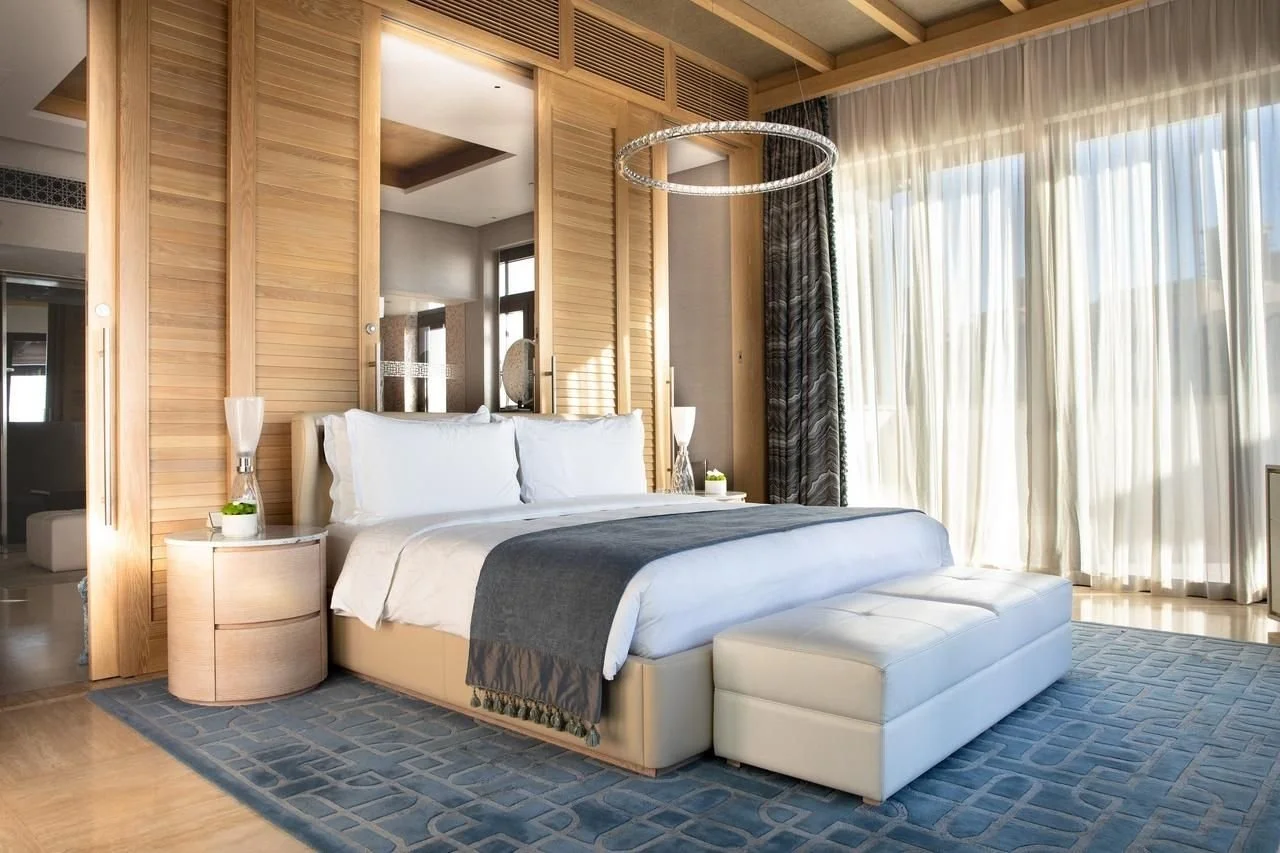 A modern bedroom with a bed, white and gray bedding, two nightstands with lamps, a large window with curtains, a wooden wall decor, and a dark area rug.