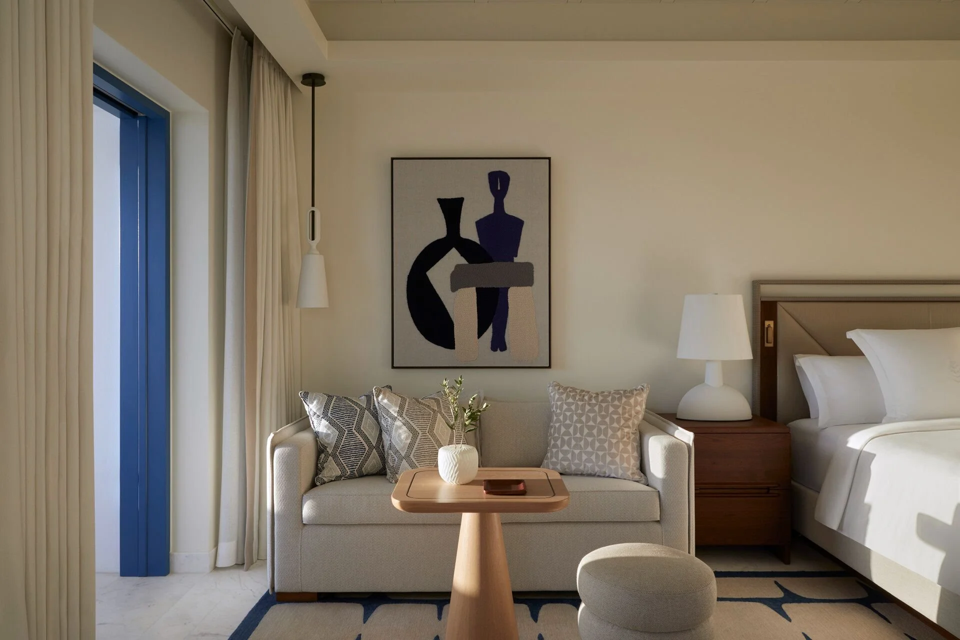 A cozy hotel room corner with a beige sofa, patterned pillows, a small wooden table with a white vase, and a painting on a wall. There is a bed with white linens and a nightstand with a white lamp. A round ottoman is in front of the sofa, and cream curtains are on a window with a blue frame.