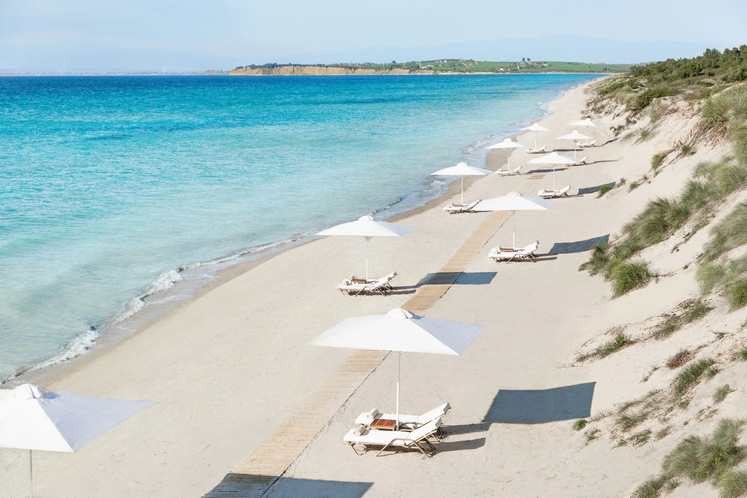 Empty beach with white umbrellas and lounge chairs along the shoreline, sandy dunes with grass on the right, turquoise water, and distant land on the horizon.