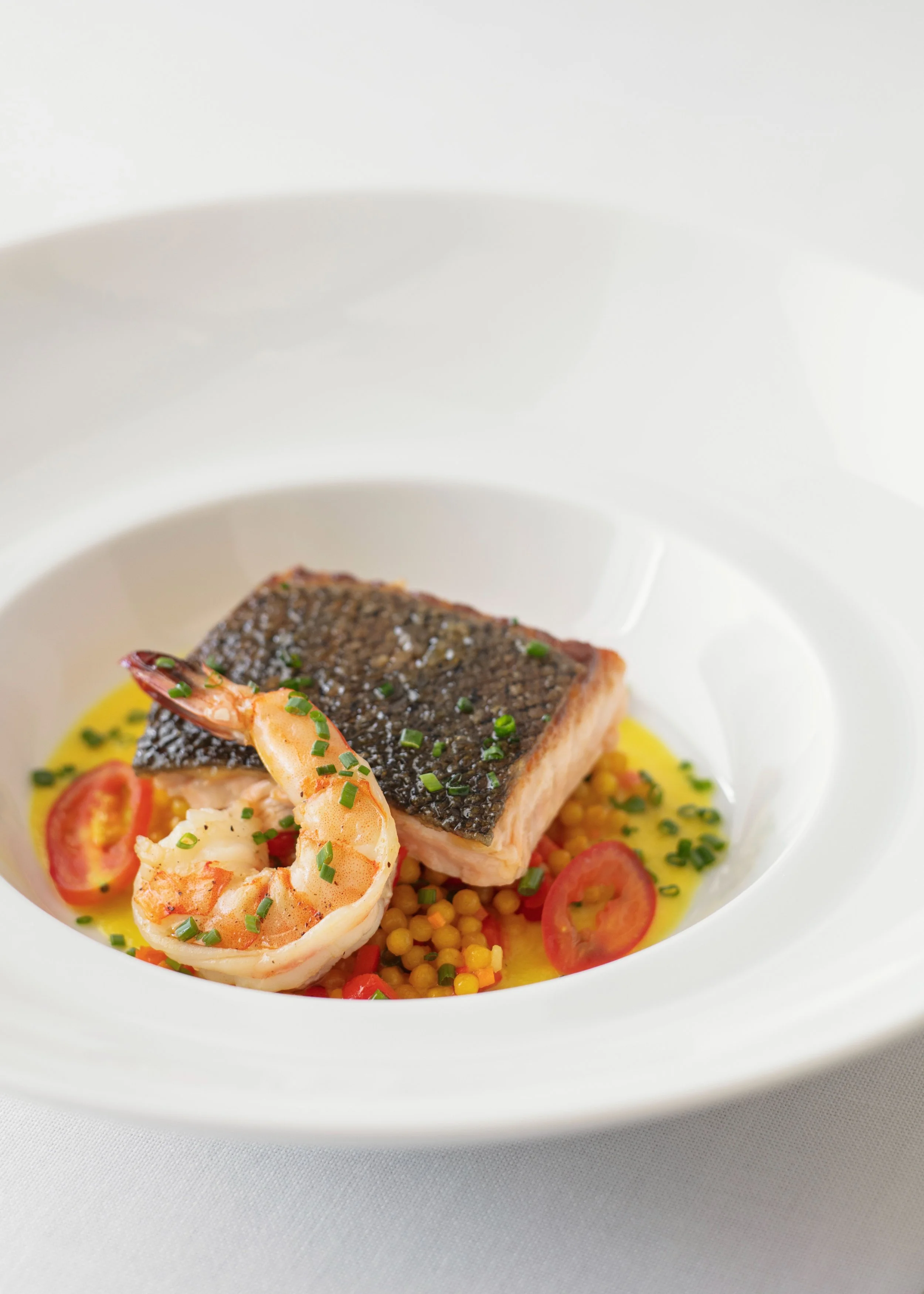 A gourmet dish featuring a piece of seared fish with crispy skin, topped with chopped chives, served with a yellow sauce, cherry tomato slices, a cooked shrimp, and a bed of small, round yellow grains on a white plate.