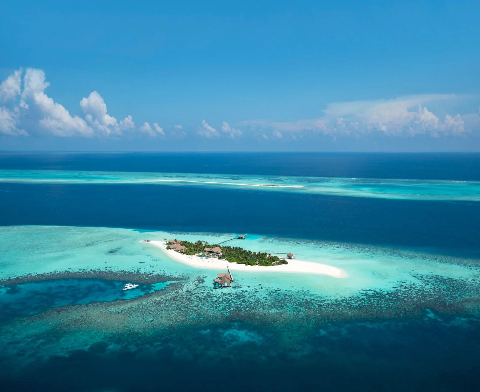 A tropical island with overwater bungalows, surrounded by turquoise water, with a boat docked near the island and an expansive blue ocean in the background.