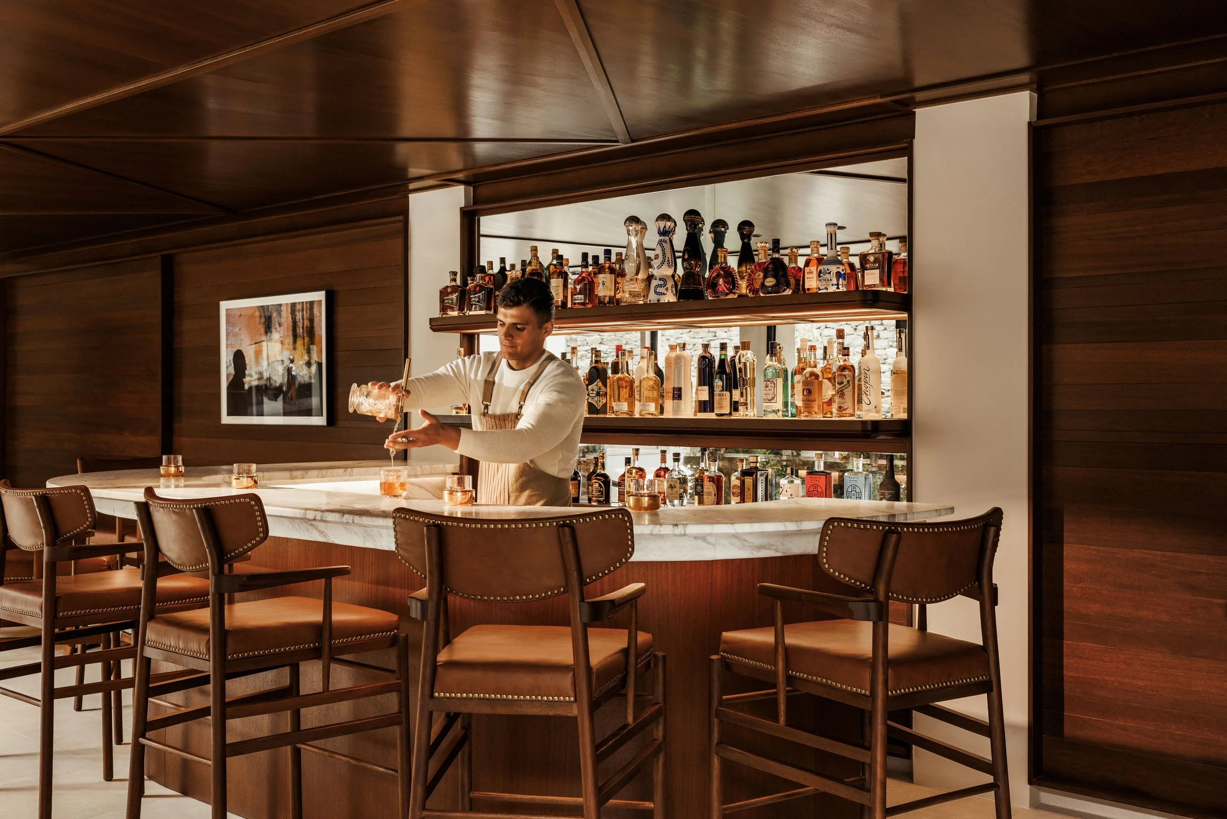 A bartender pouring a drink at a marble bar top in a room with wooden paneling and a display of assorted liquor bottles on shelves behind him.