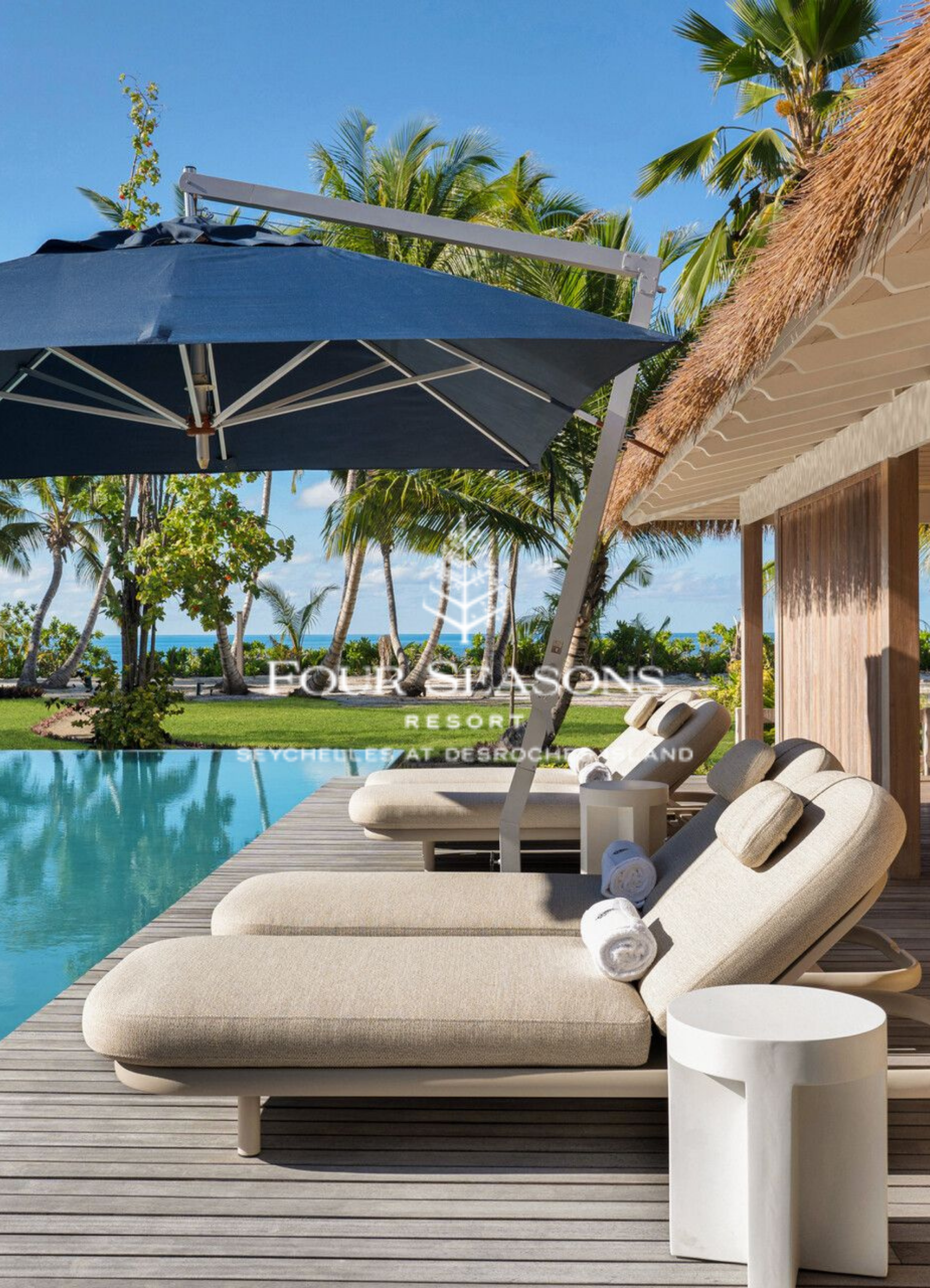 Poolside view with beige lounge chair and rolled towels, white side table, pool, tropical trees, thatched roof structure, and clear blue sky at Follyshon Resort in Seychelles.