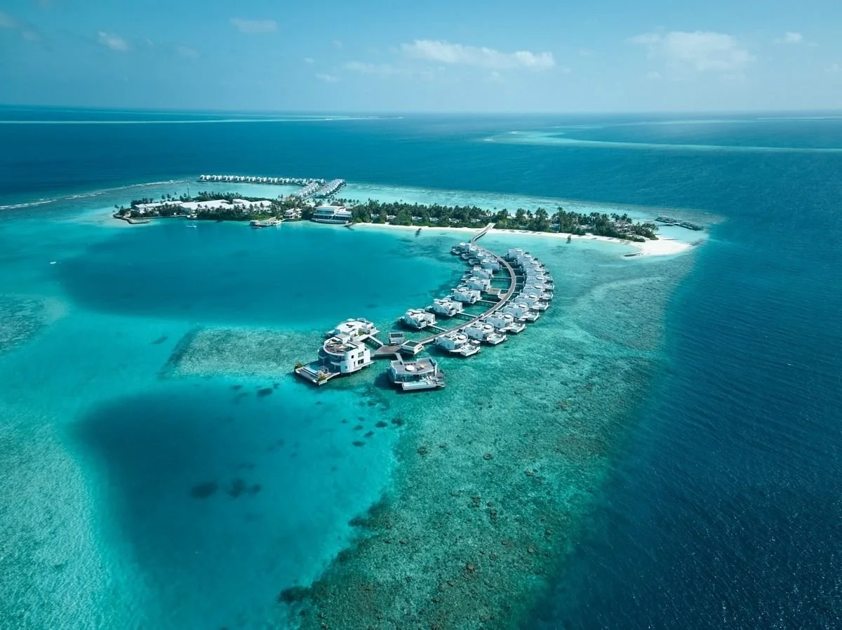 Aerial view of an overwater bungalow resort on a tropical island with clear turquoise waters and coral reefs.