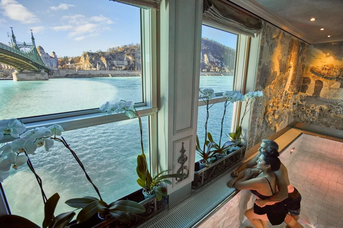 Two women sitting at the edge of a hot tub by large windows overlooking a river, with a cityscape and bridge outside, decorated with white orchids inside.