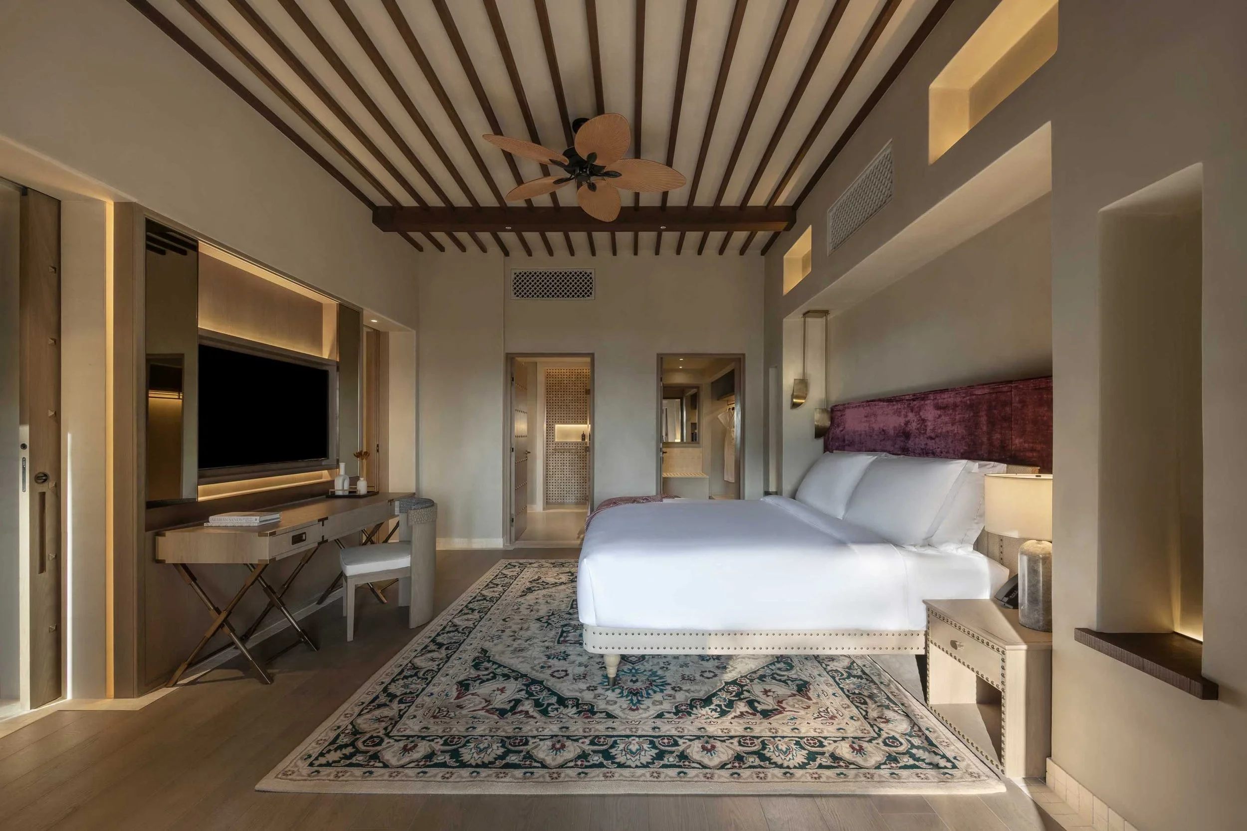 A modern hotel room with a large bed, a flat-screen TV, a desk and chair, a ceiling fan, and decorative lighting on the walls and ceiling.