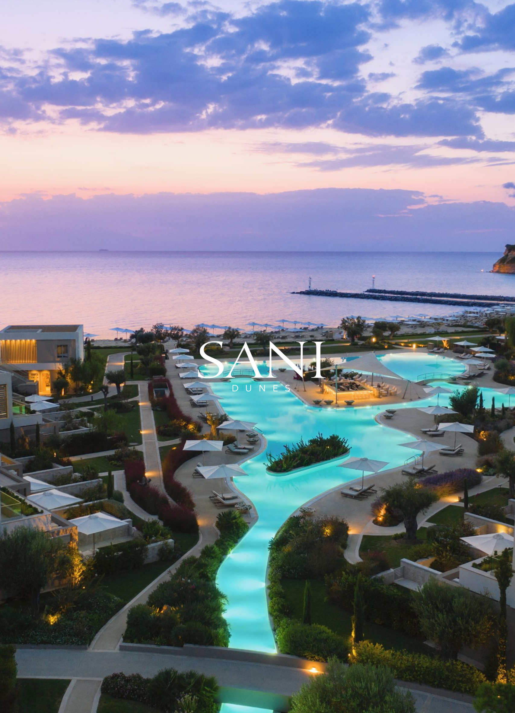 Overlooking the pool area at sunset or dusk, with the ocean and horizon in the background. The pool is lit up with turquoise lighting, surrounded by lounge chairs, umbrellas, and landscaped greenery. The ocean has calm waters, with a breakwater and a lighthouse or navigation point at the pier. The sky is partly cloudy with shades of purple and pink.