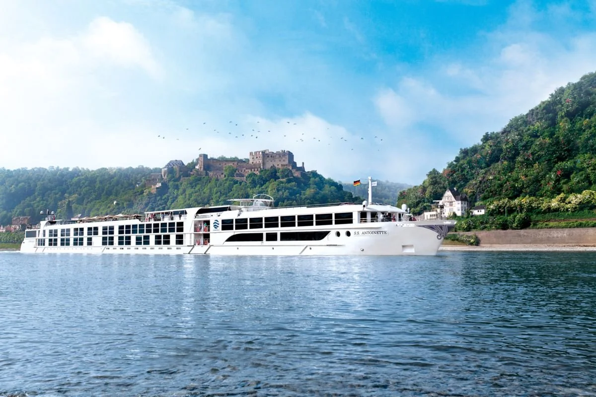 A large white cruise ship sailing on a river with green hills and a castle in the background under a partly cloudy sky.