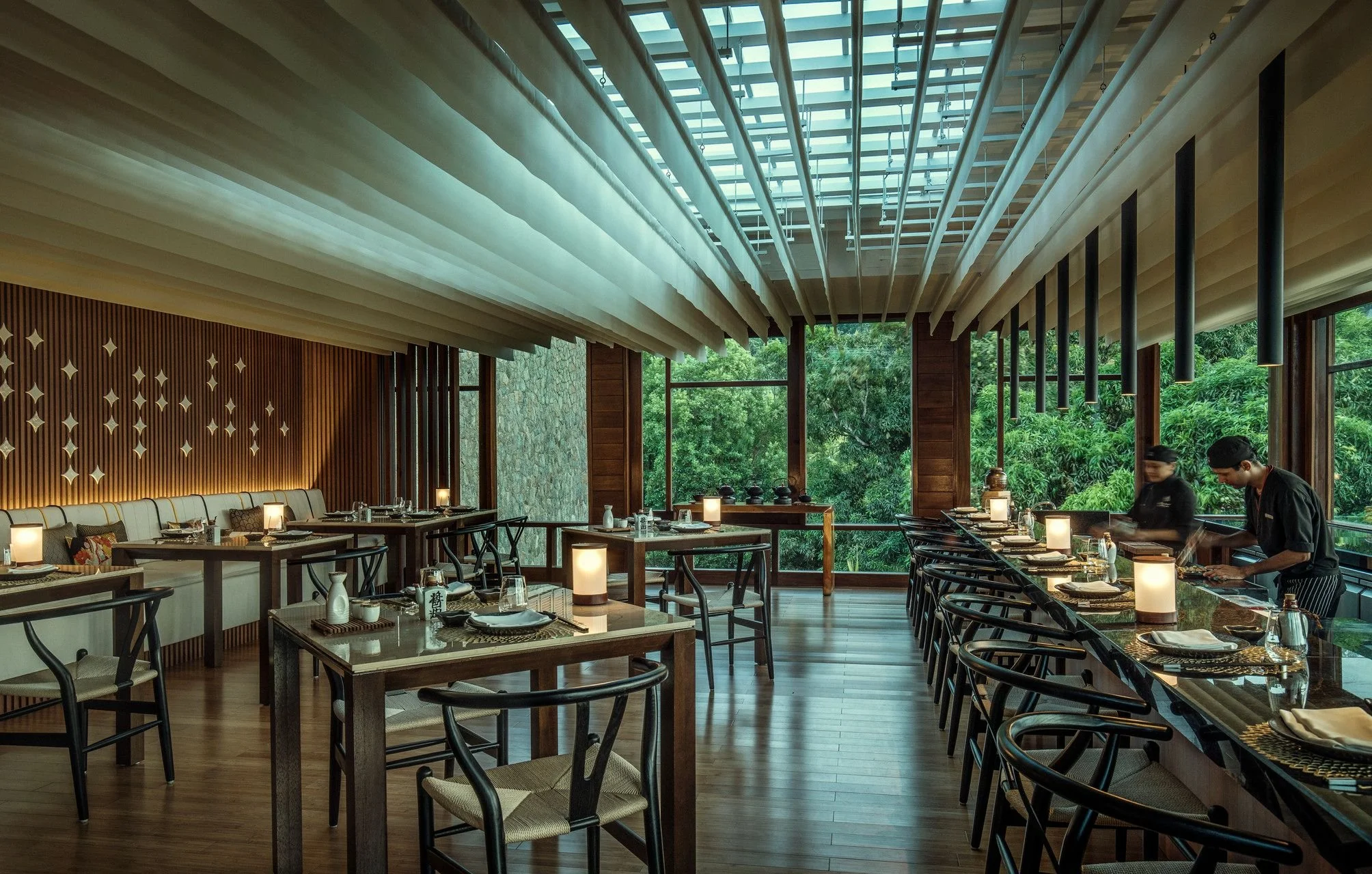 Modern restaurant interior with wooden floors, large windows overlooking green trees, and tables set for dining with candles and tableware. Staff members are preparing food at the right side.