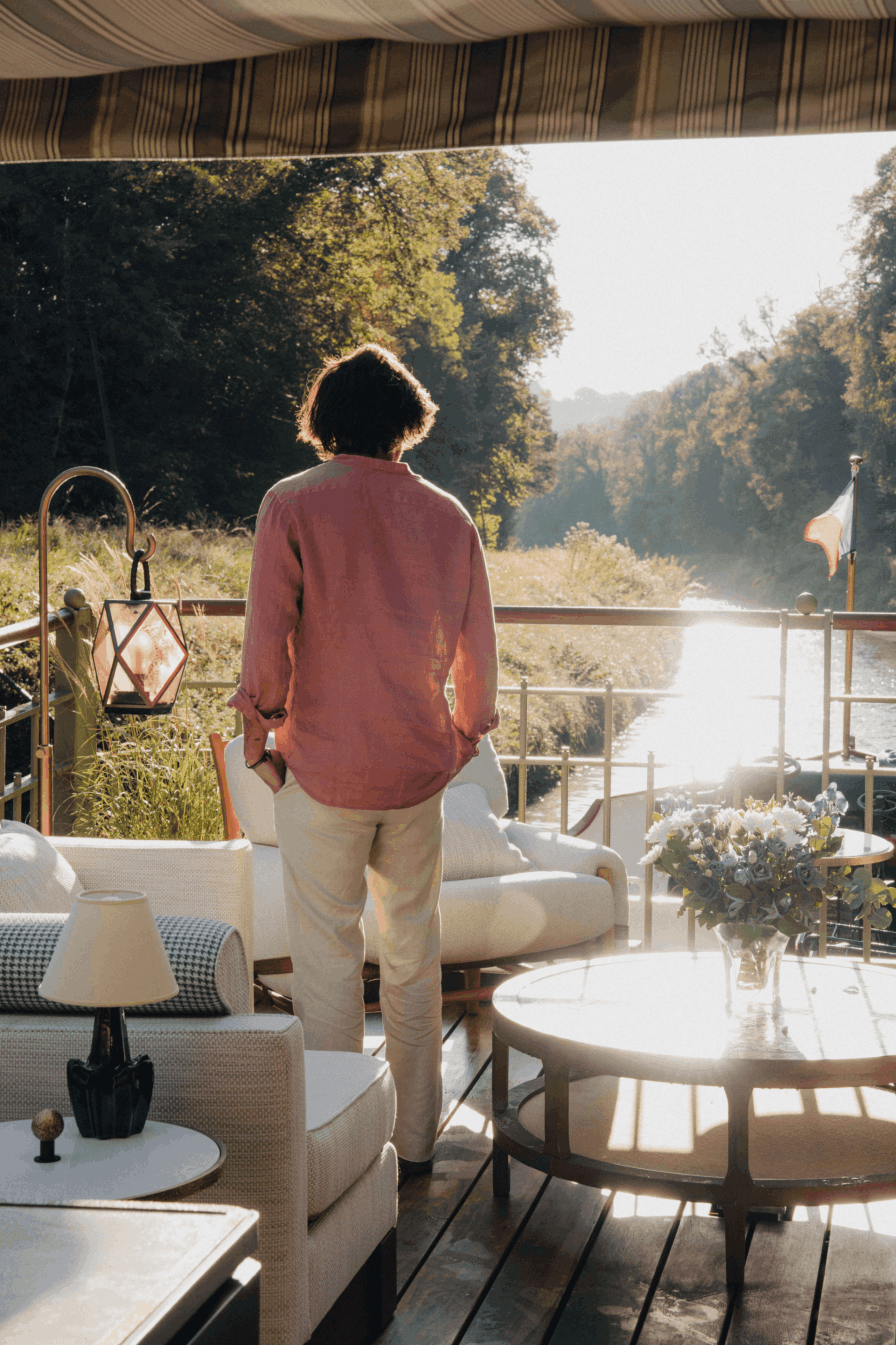 Person with short dark hair wearing a pink blouse and beige pants, standing in a sunlit airy living space overlooking a river and lush trees, with a striped awning above and floral arrangement on a side table.