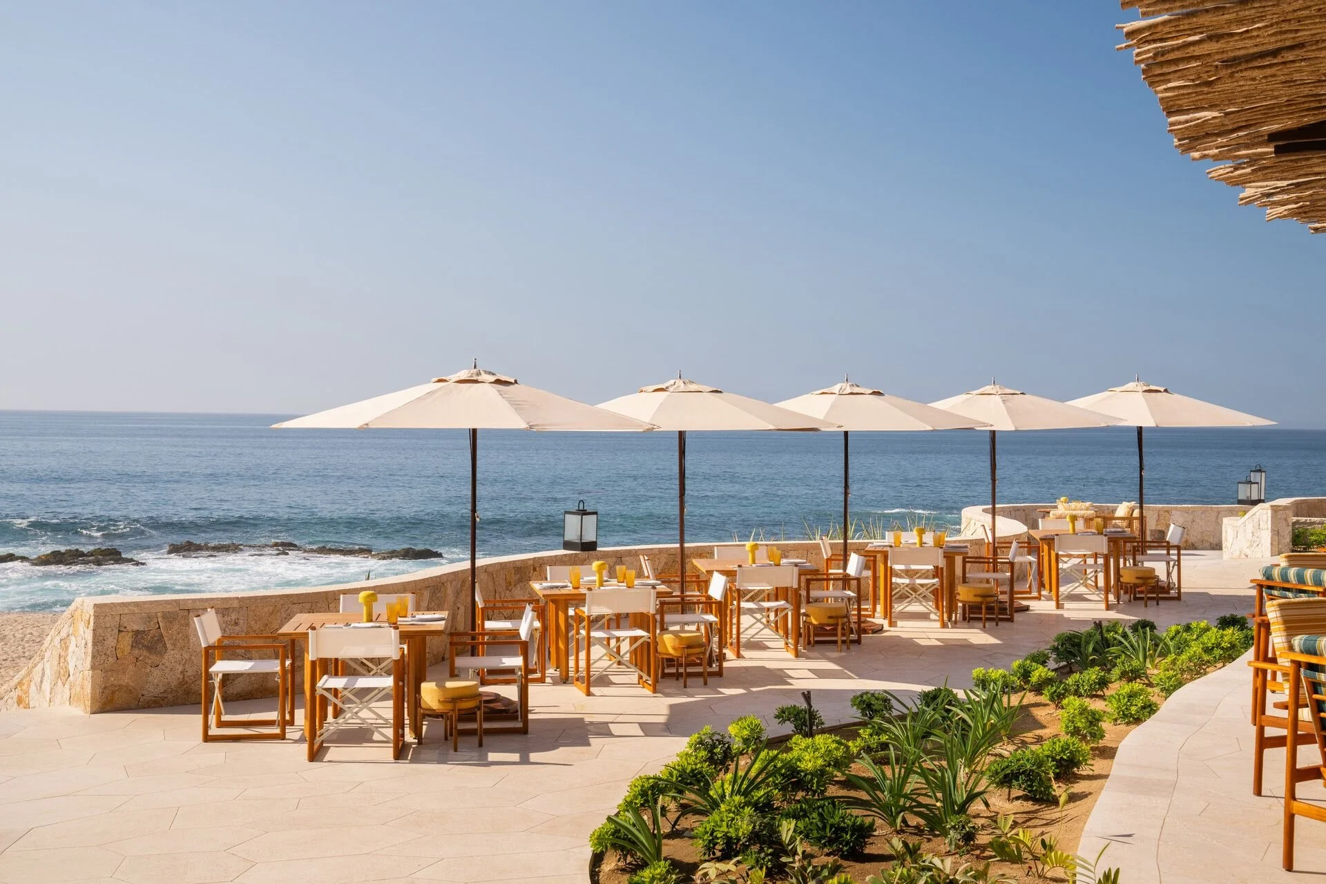 Outdoor seating area with tables, chairs, and umbrellas overlooking the ocean at a beachside restaurant or bar.