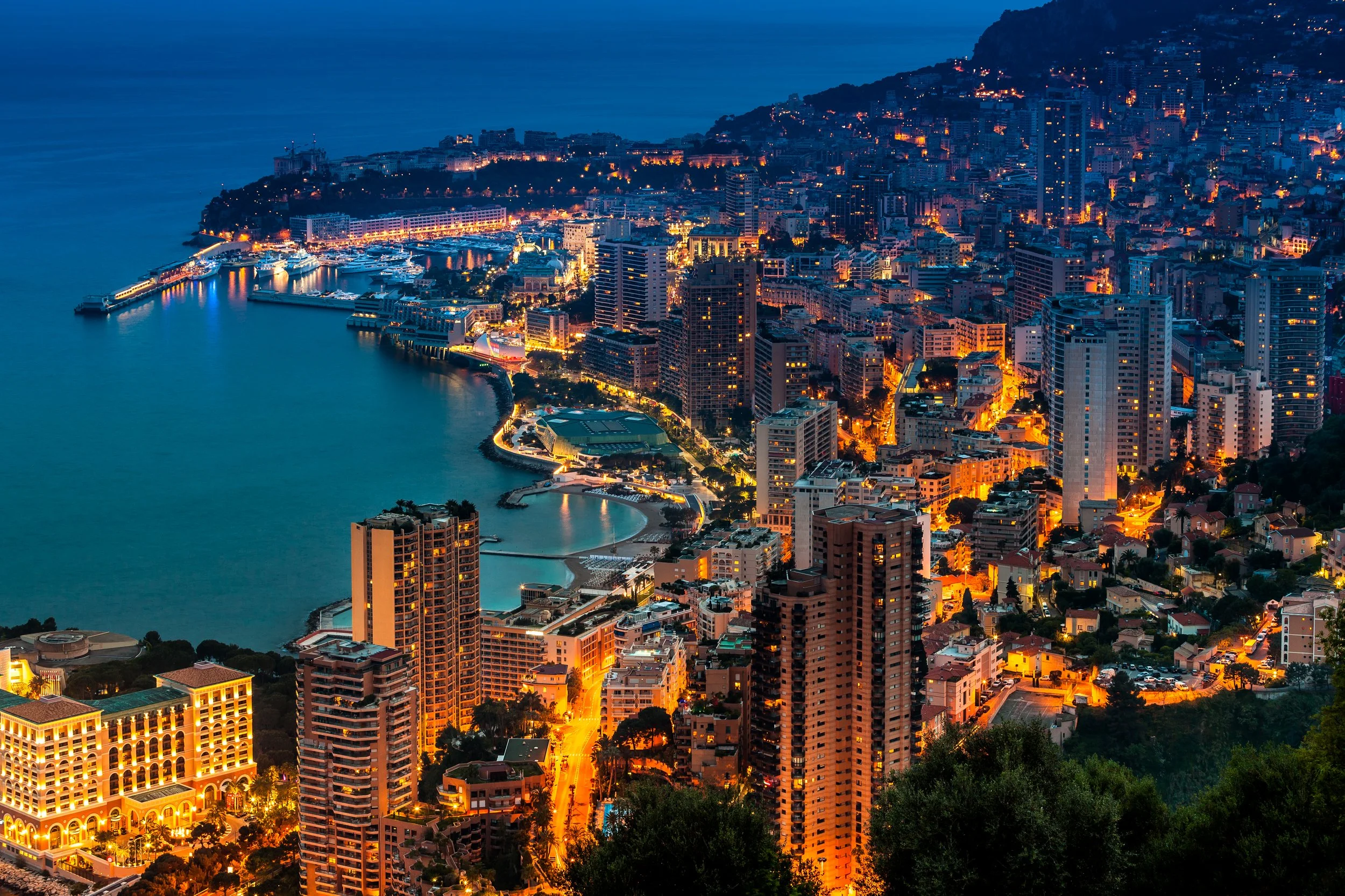 Night view of a waterfront city with high-rise buildings illuminated, a harbor with boats docked, surrounded by a hillside with houses, and city streets glowing with streetlights.