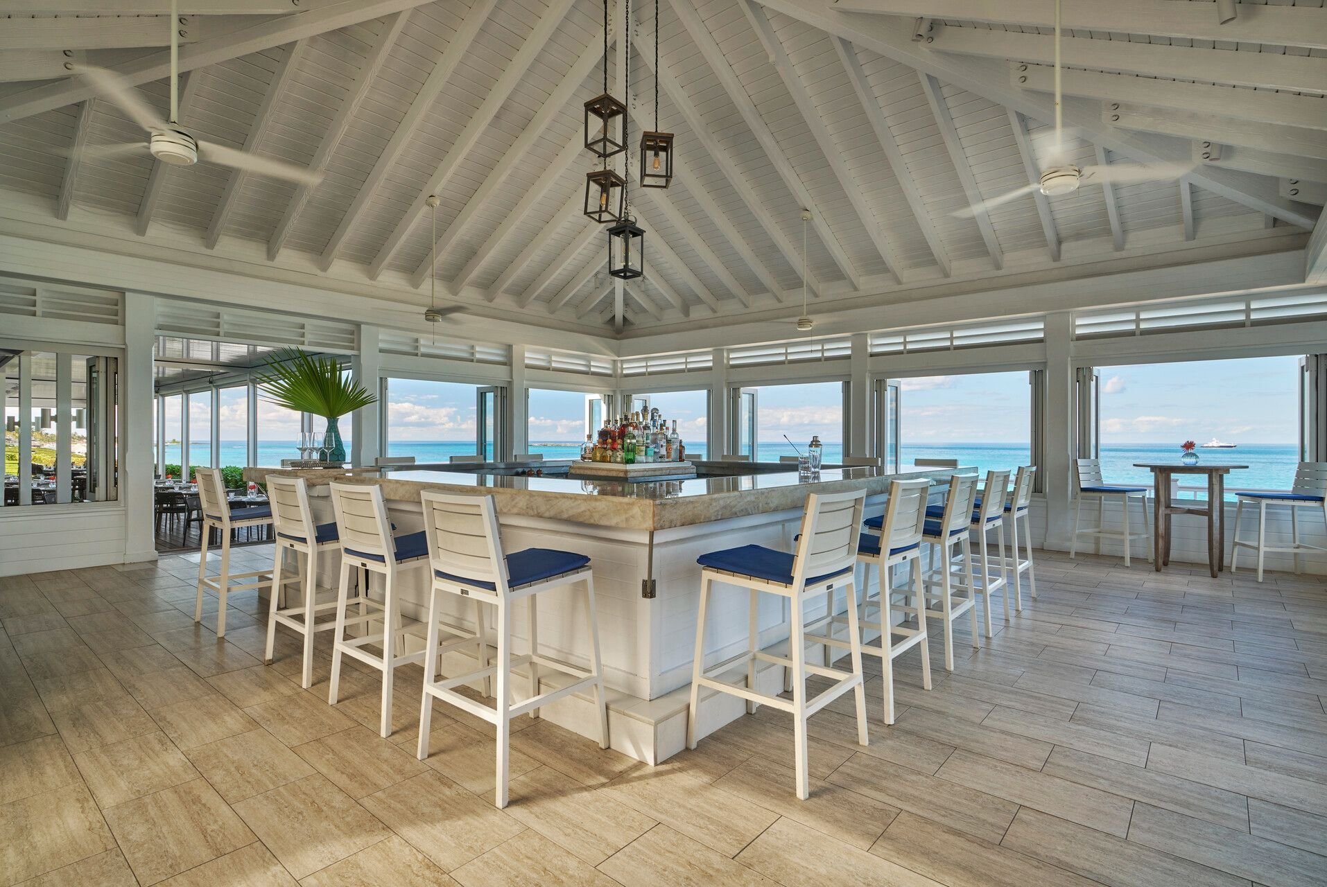 An open-air beach bar with a white wooden ceiling and chandeliers, overlooking the ocean, featuring a large marble-topped bar with barstools and a view of the sea through open windows.