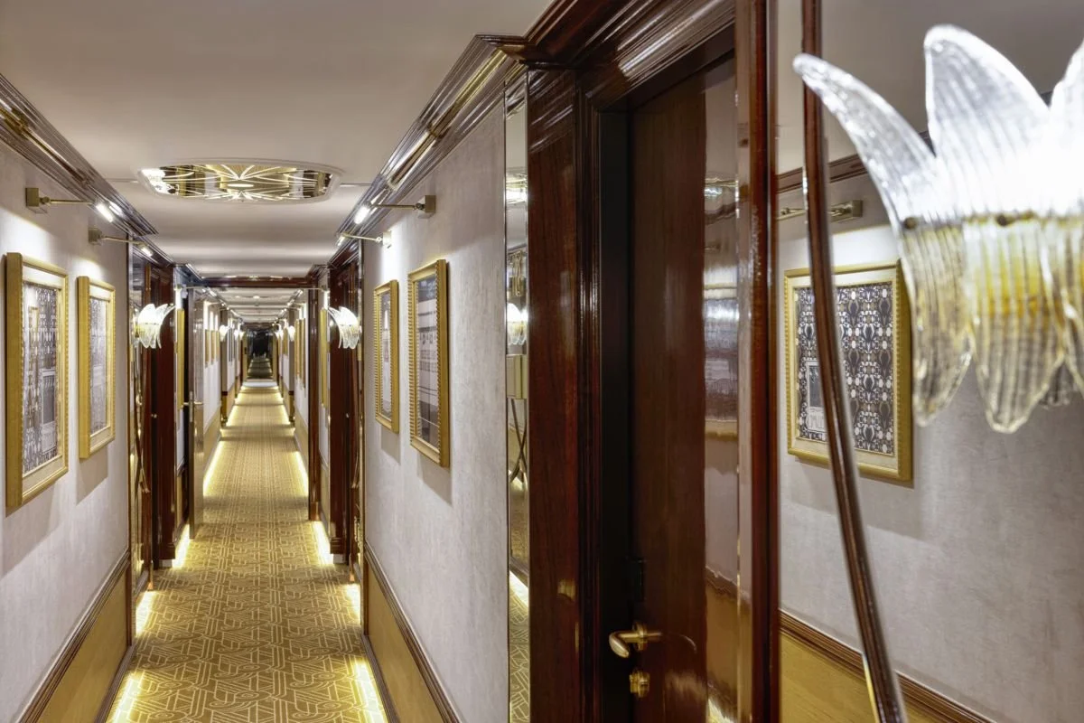 Long hotel corridor with framed pictures on cream-colored walls, gold-colored lighting fixtures, and a patterned yellow carpet.