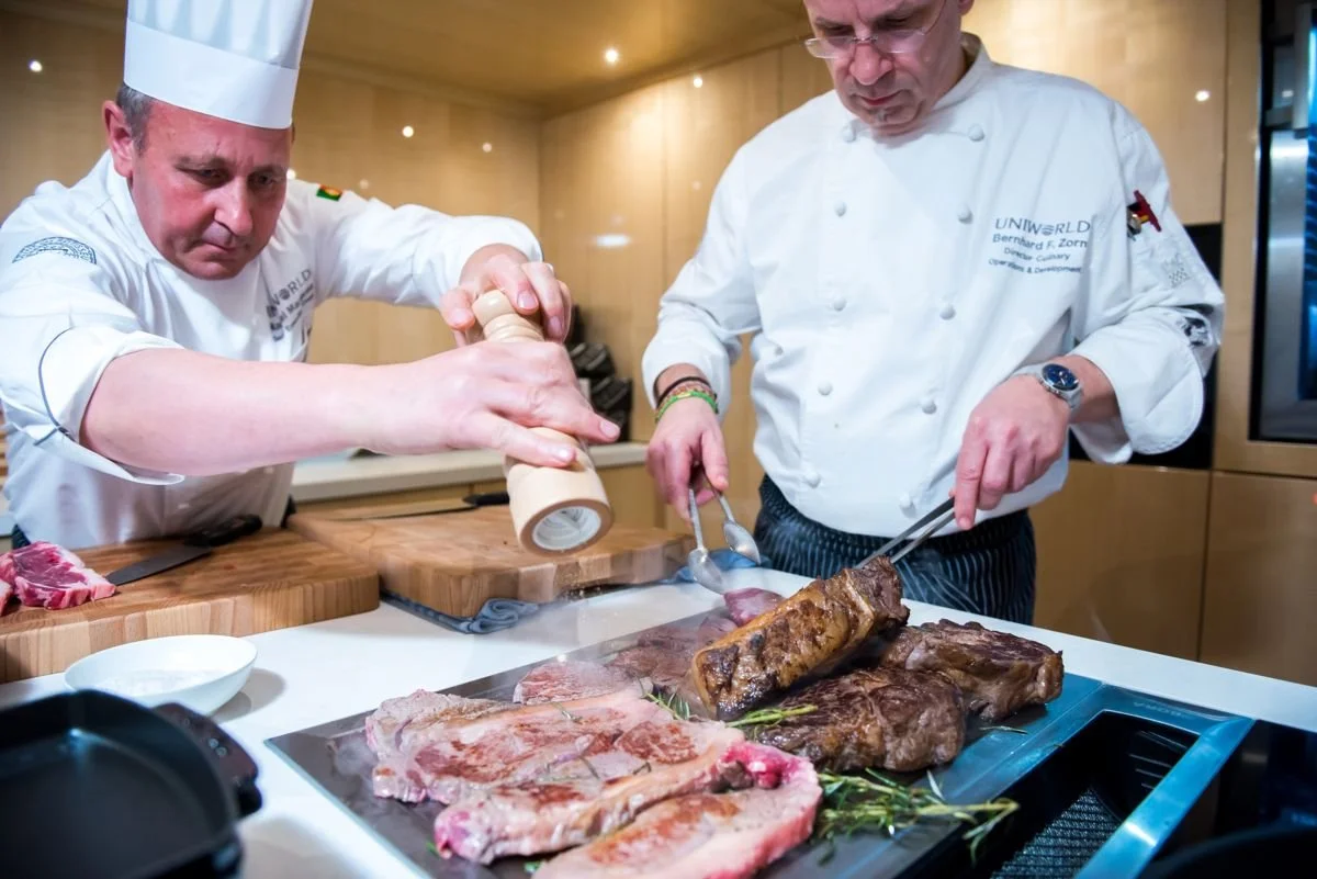 Two chefs seasoning and preparing large cuts of beef and ribs on a kitchen counter.